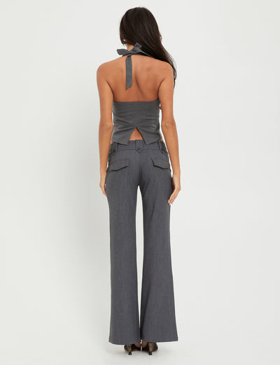 A woman with long dark hair stands facing away from the camera, wearing the stylish TRISTAN VEST - GREY from TIGERMIST. The vest features an open back and a bow at the neck, paired with matching high-waisted, wide-leg pants. The background is plain white.