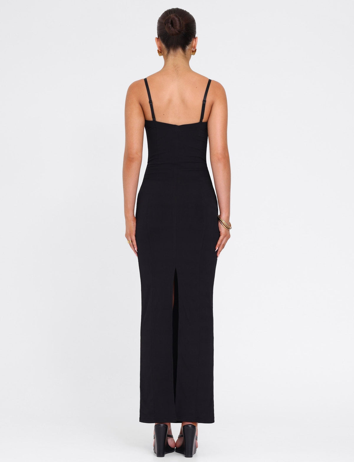 A woman faces away in the TIGERMIST ALLURA MAXI DRESS - BLACK, a fitted bodycon style featuring thin straps, lace bra cups, and a back slit. She pairs it with black heels and a bracelet, her hair in a bun against a plain white background.