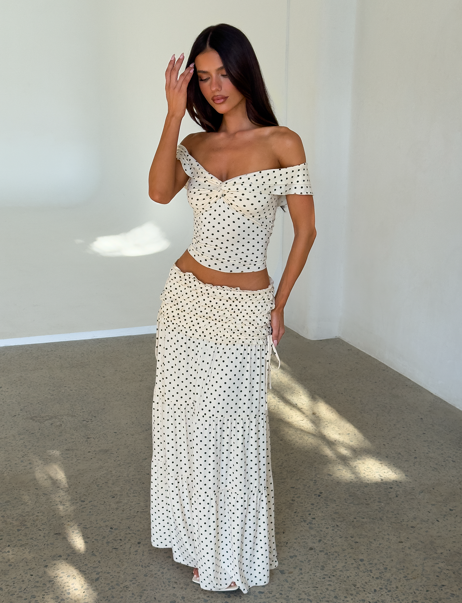 A woman stands indoors in the TIGERMIST DENALI TOP - CREAM DOT, an off-shoulder top with black polka dots, paired with a matching long skirt. She has long dark hair and touches her forehead with one hand.