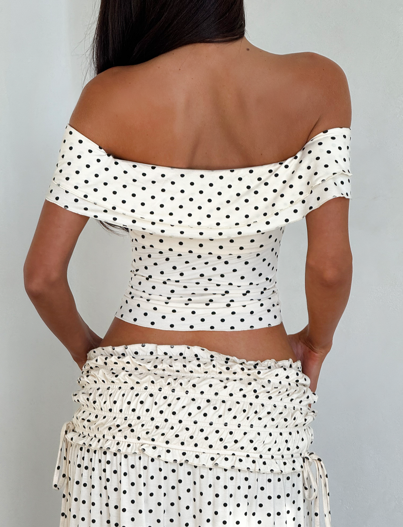 A woman with loose dark hair stands against a plain light background wearing the TIGERMIST DENALI MAXI SKIRT - CREAM DOT and a matching off-the-shoulder, white polka dot top.