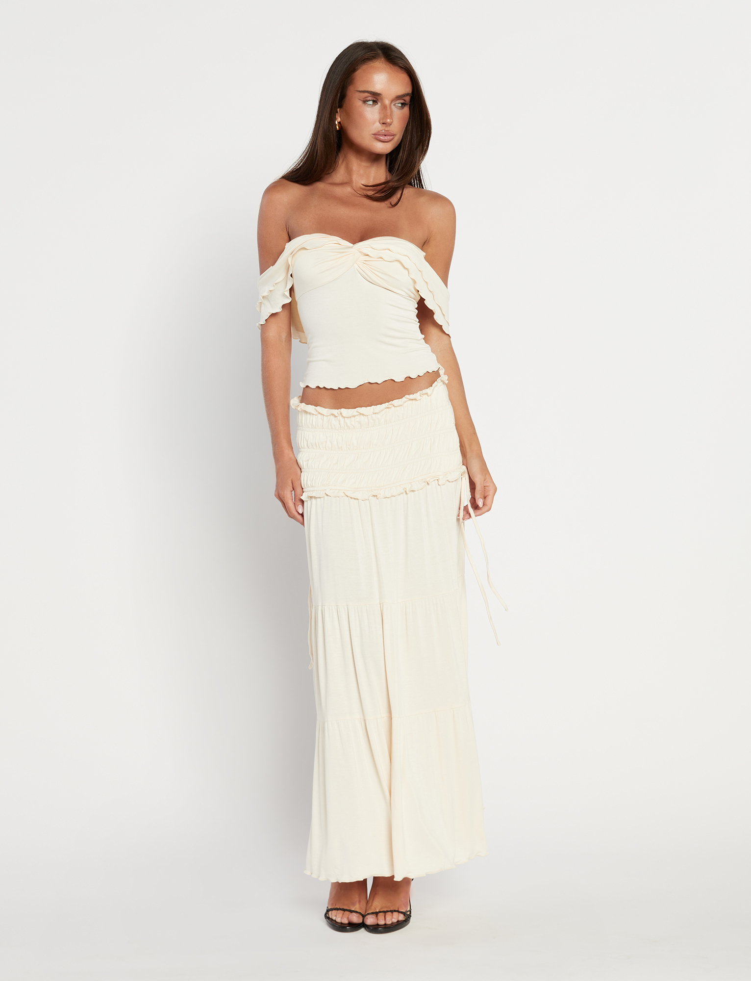 A woman with long brown hair wears the TIGERMIST DENALI MAXI SKIRT in Neutral: Bone, paired with an off-shoulder cream ruffled crop top, standing against a white background and styled with black heeled sandals.