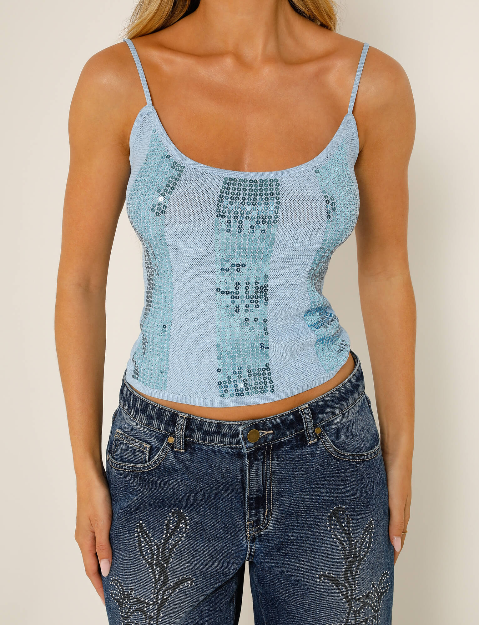 A person wearing the JUNNO TOP - BLUE from TIGERMIST, paired with blue jeans featuring embroidered detailing on the thighs, stands against a neutral background.