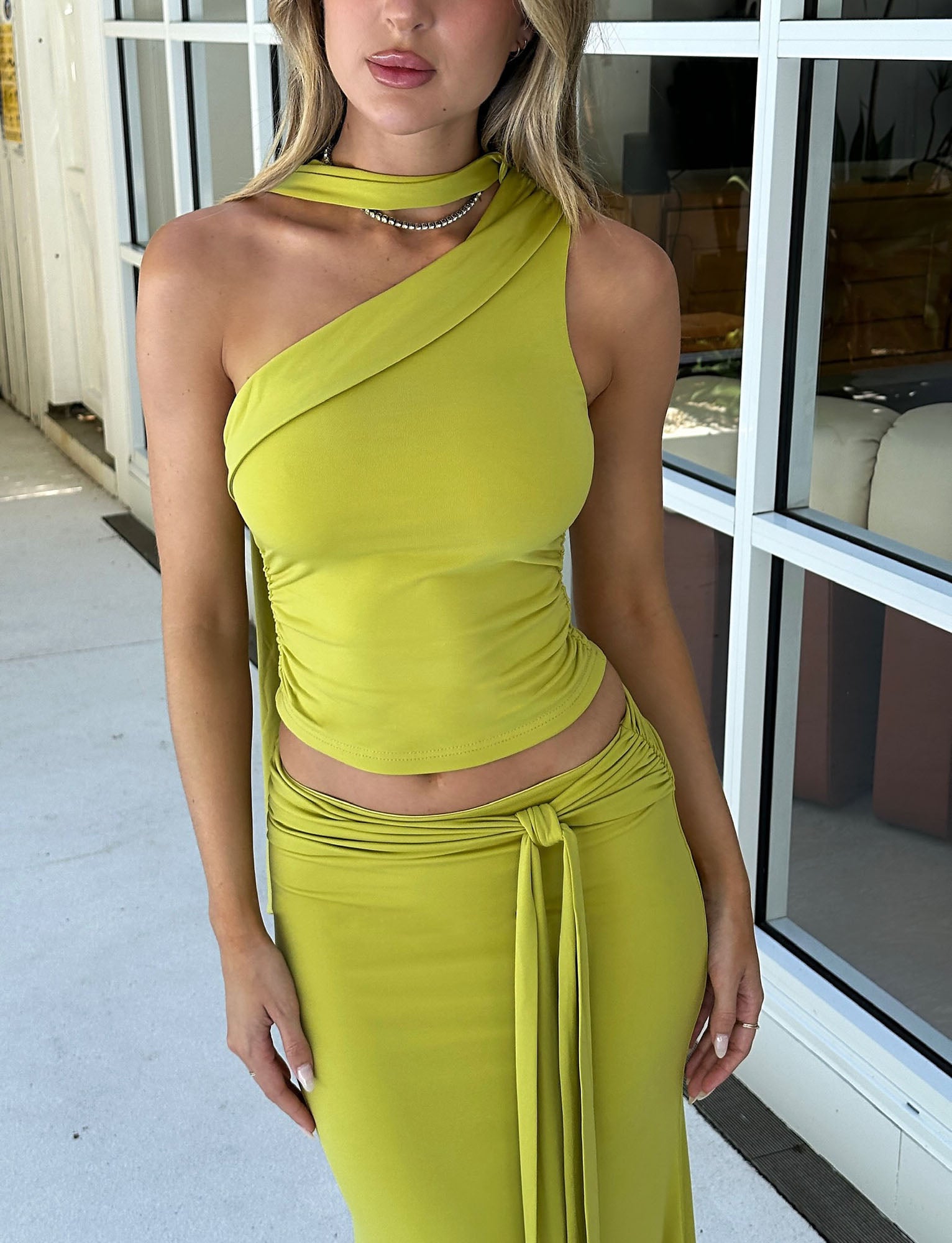 A person is shown wearing the ALLIRA TOP in Chartreuse from TIGERMIST, along with a matching skirt featuring a front tie. They are standing in front of a large window and white wall. Their hair is styled straight, and they are accessorized with a necklace.
