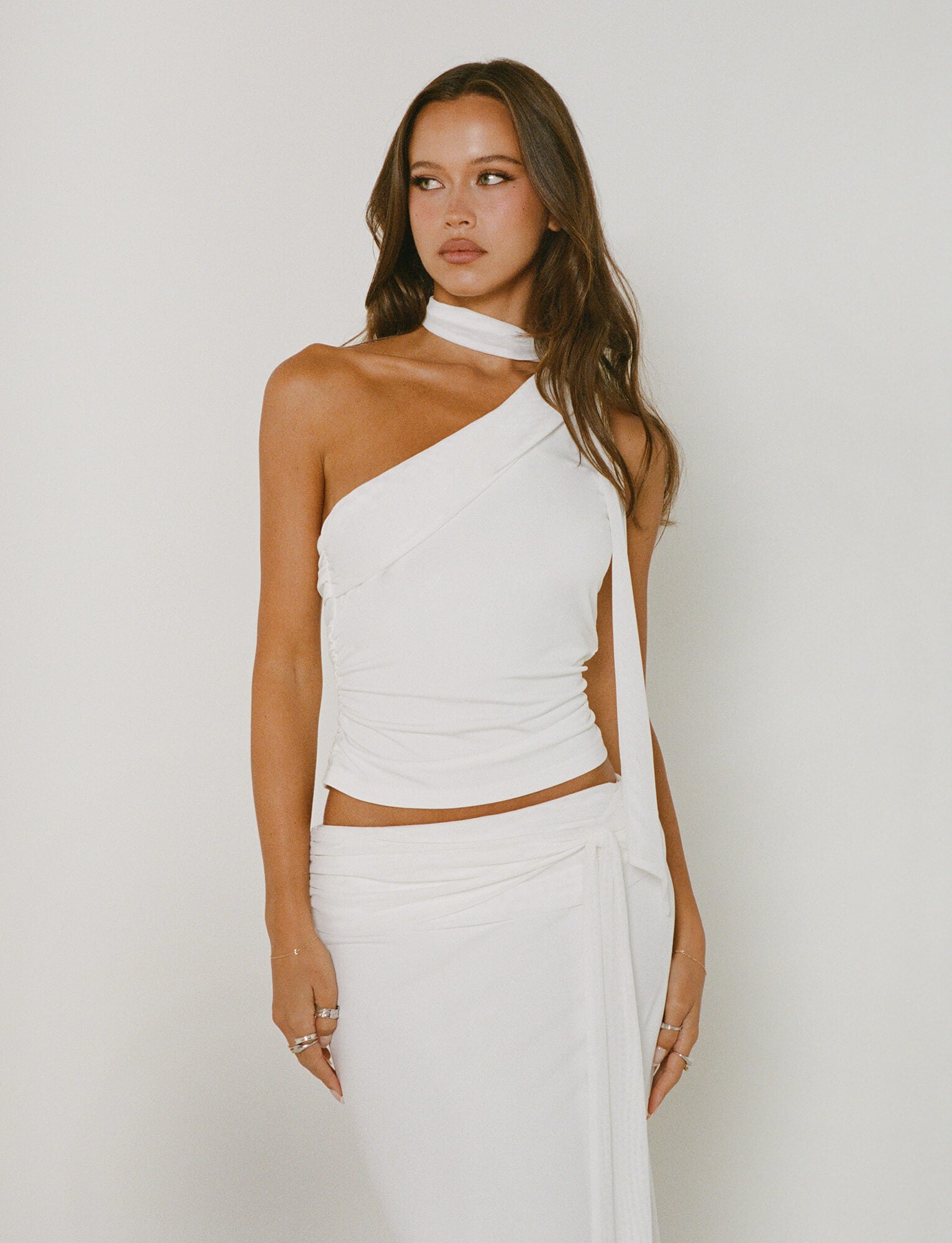 A woman with long, wavy brown hair is wearing the ALLIRA TOP - WHITE and a matching skirt from TIGERMIST. She stands against a plain background, looking to the side. Her look is completed with rings on her fingers.