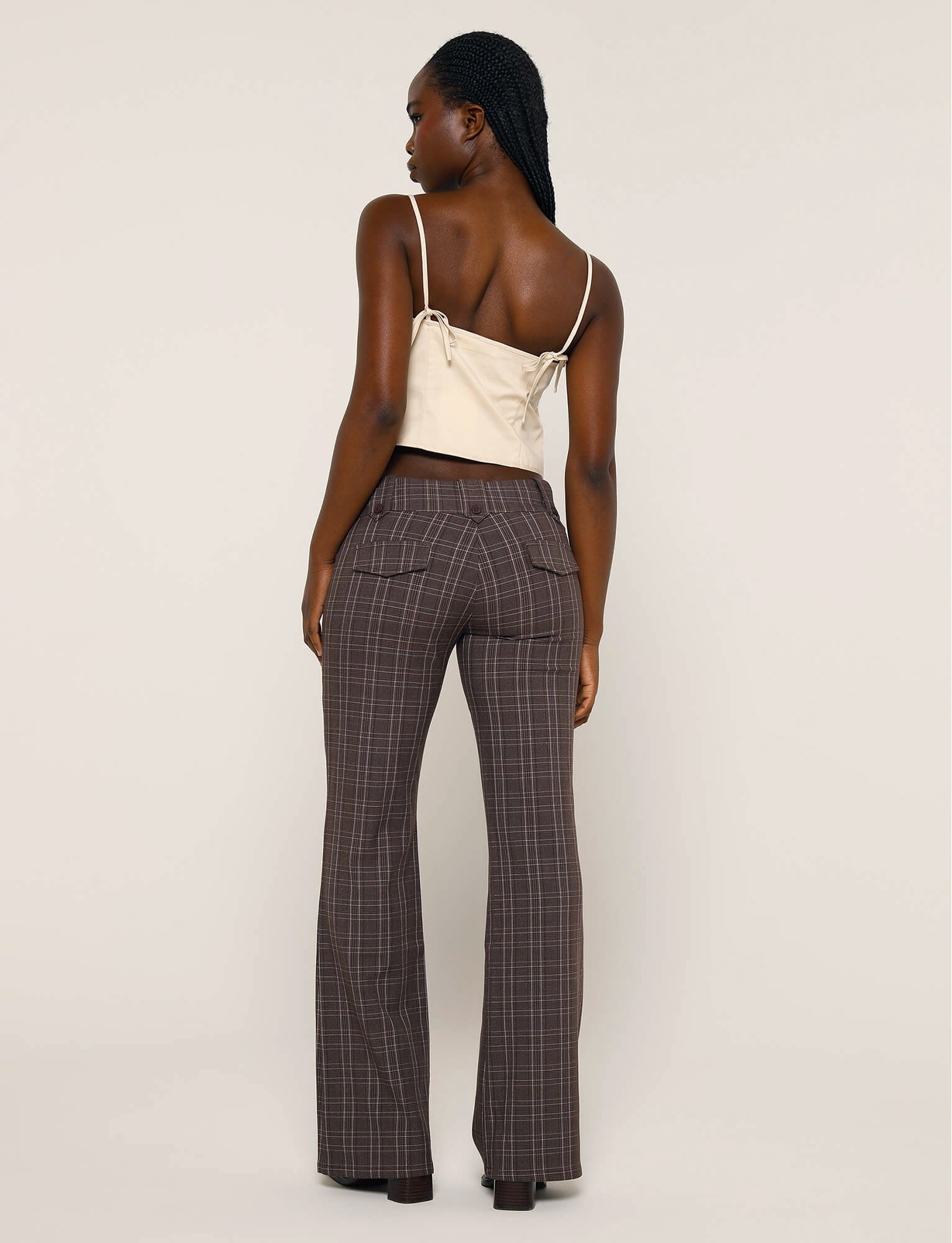 A person with braids is seen from behind, wearing a sleeveless beige top with tie details on the shoulders and the KITTIE PANT - BROWN : CHOCOLATE CHECK from TIGERMIST. The backdrop is neutral.