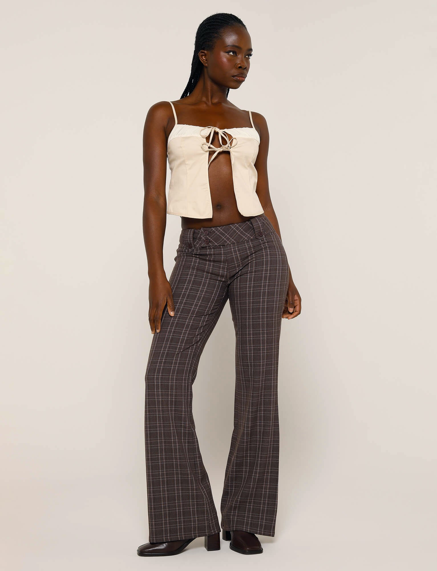 A woman stands confidently against a plain background, wearing a beige, sleeveless crop top with a front tie detail and the KITTIE PANT - BROWN : CHOCOLATE CHECK from TIGERMIST. She completes the look with black ankle boots.