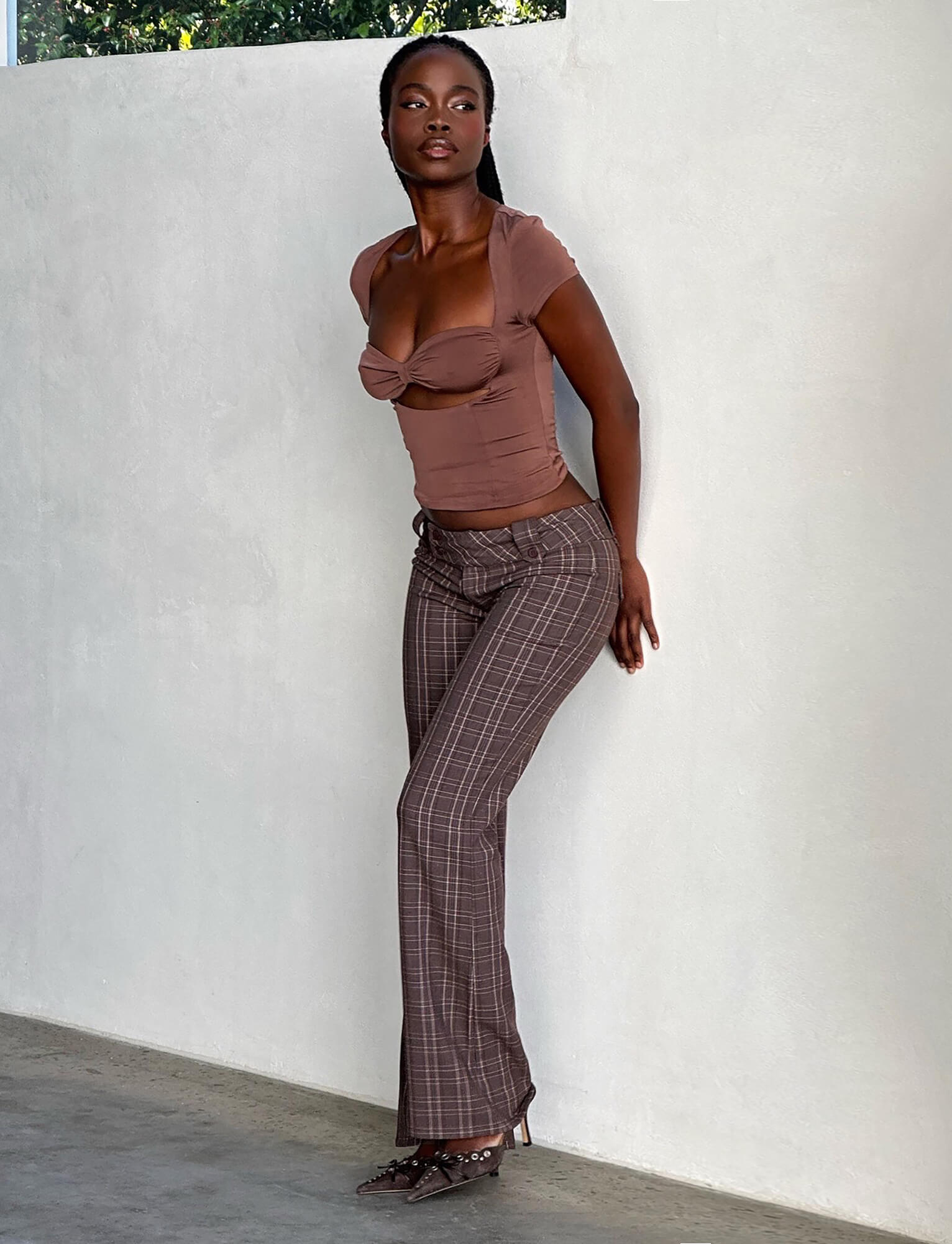 A woman stands against a white wall, exuding confidence in her TIGERMIST ensemble. She wears a fitted brown top with short sleeves and a knot detail at the chest, paired with the TIGERMIST KITTIE PANT - BROWN : CHOCOLATE CHECK and matching heeled shoes. She gazes off to the side, her expression radiating assurance.