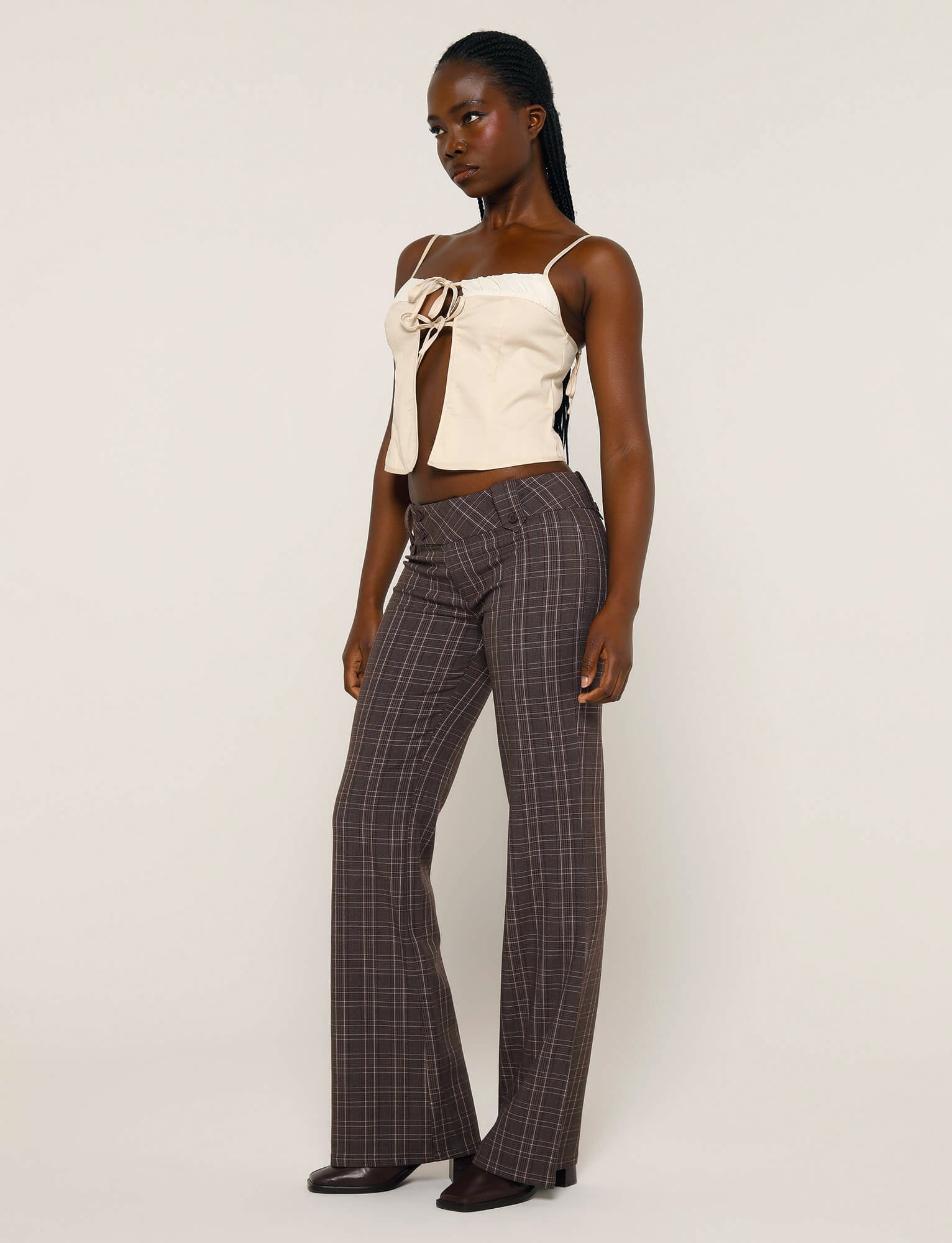A woman stands against a plain background, wearing a beige sleeveless top with a front tie detail and the KITTIE PANT - BROWN : CHOCOLATE CHECK from TIGERMIST. She has long dark braided hair and is wearing dark brown shoes. She gazes slightly to her left.