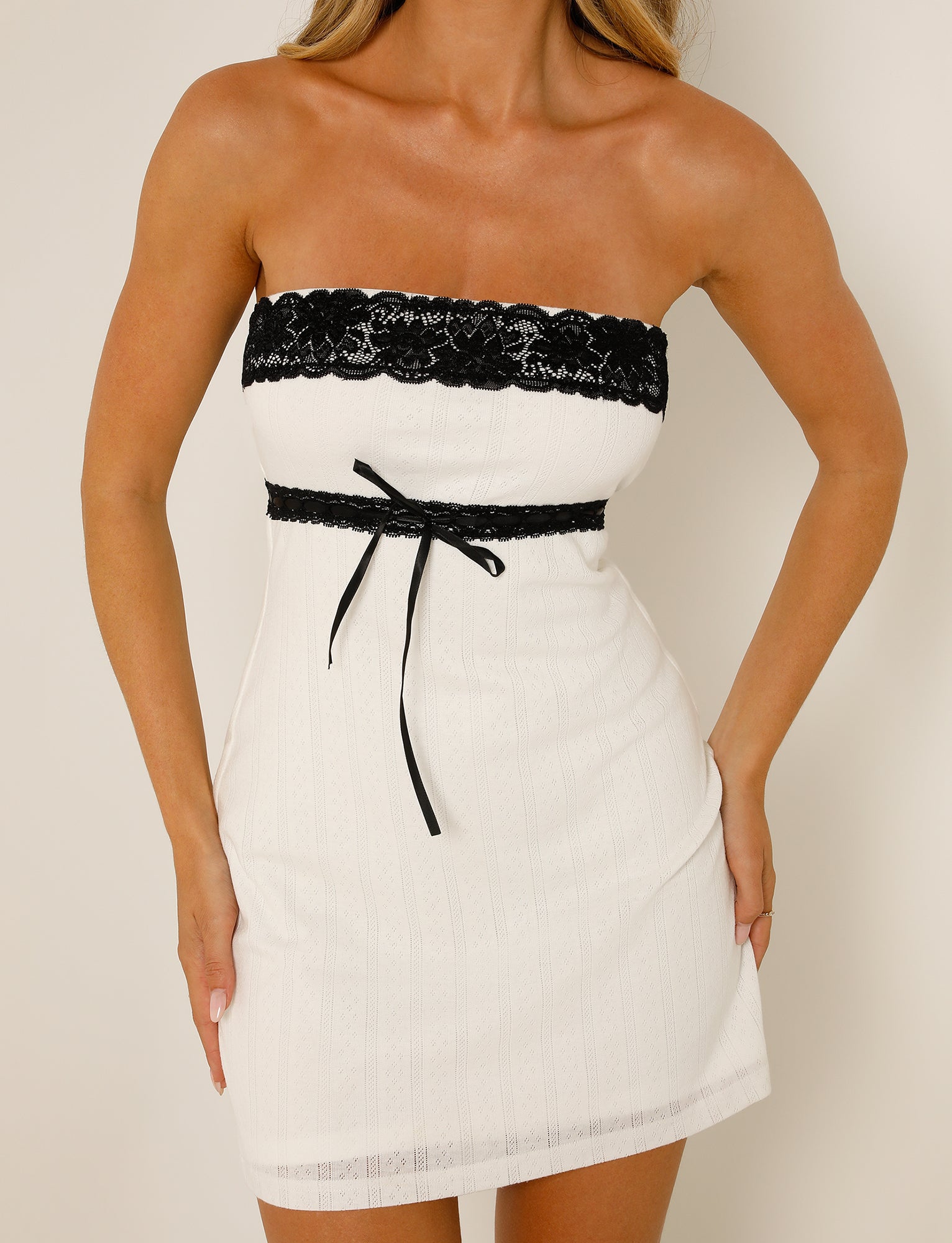 A woman is seen in the TIGERMIST MARCIA MINI DRESS - WHITE, a stylish strapless outfit featuring intricate black lace detailing along the top and a chic black ribbon tied around her waist. The dress falls above the knee, and she poses with her hands resting on her hips against a plain white background.
