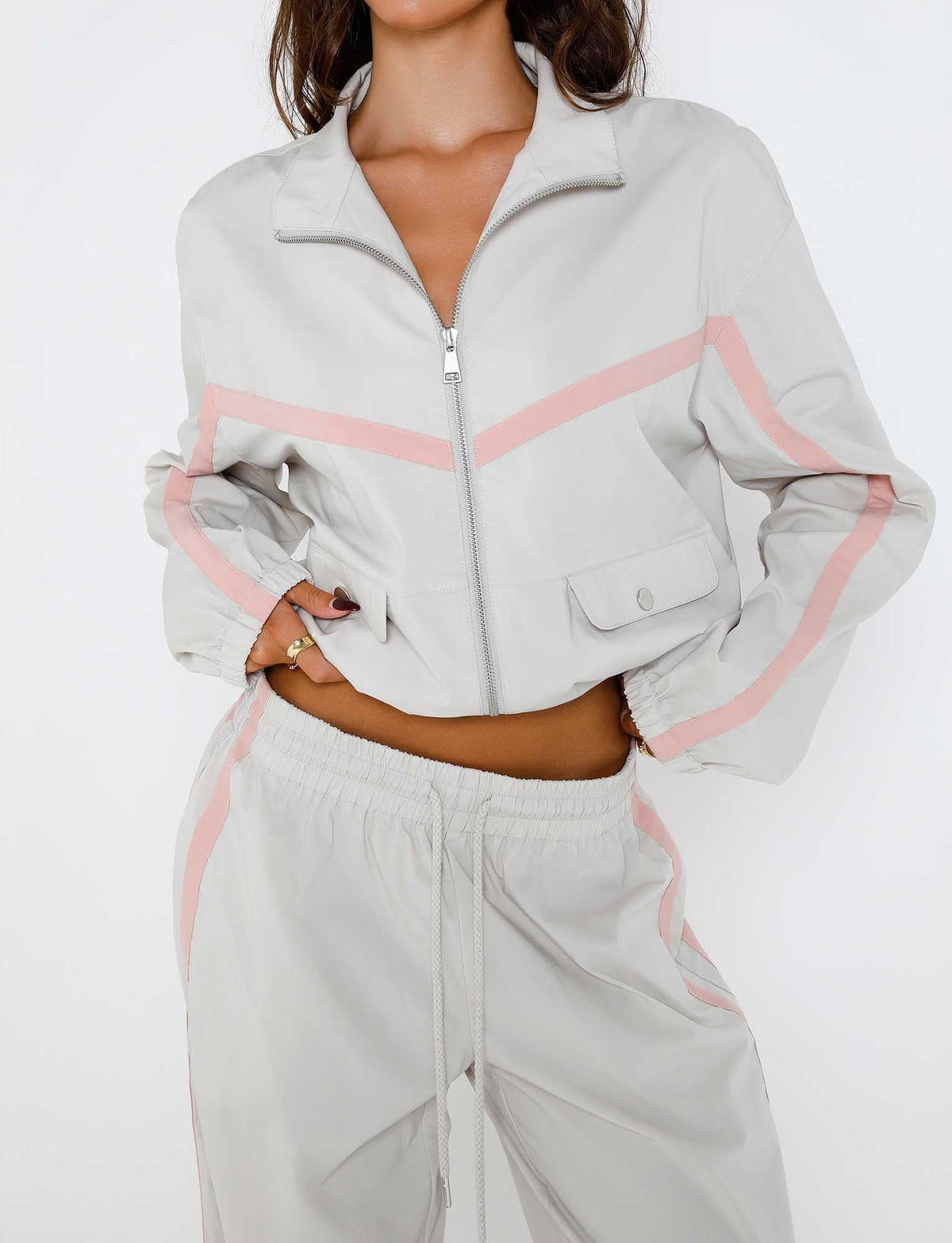 A person wearing the TIGERMIST KAHANNA JACKET - GREY : DOVE GREY with pink accents and matching light gray drawstring pants. The jacket features two front pockets, and the person's hands are resting on their hips. The background is plain white.