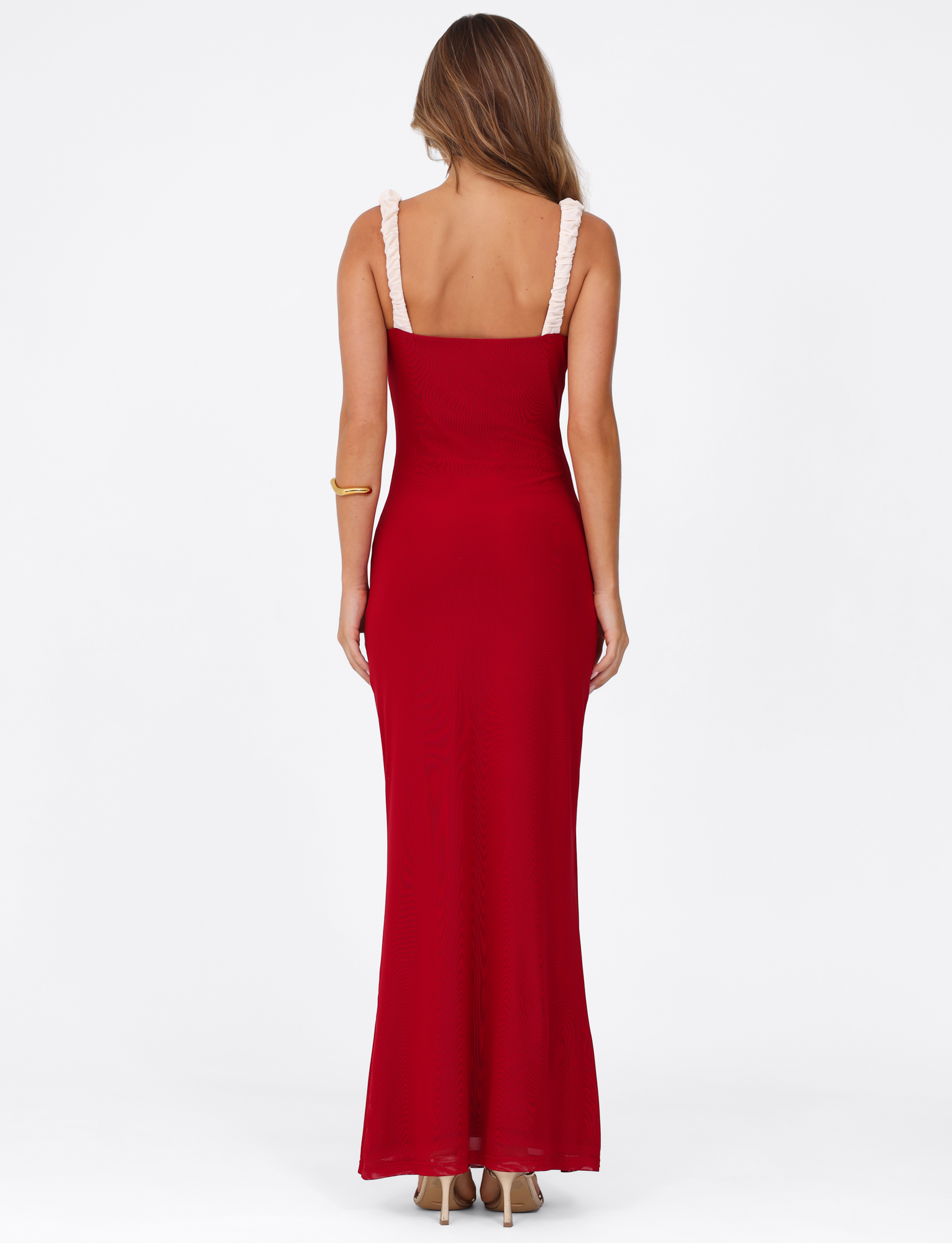 A woman with light brown hair and beige heels faces away, wearing the TIGERMIST AMARAN MAXI DRESS - RED: BURGUNDY with wide cream straps and a gold bracelet on her left arm, set against a plain white background.