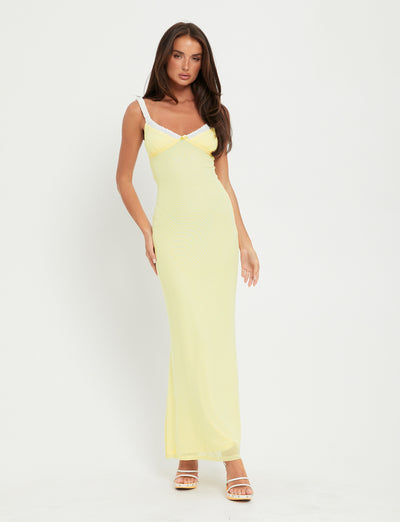 A woman stands against a plain white background, wearing the AMARAN DRESS - YELLOW : LEMON from TIGERMIST, a fitted, long yellow dress with thin white straps and a V-neckline. She has long, straight brown hair and is also wearing white strappy high-heeled sandals.