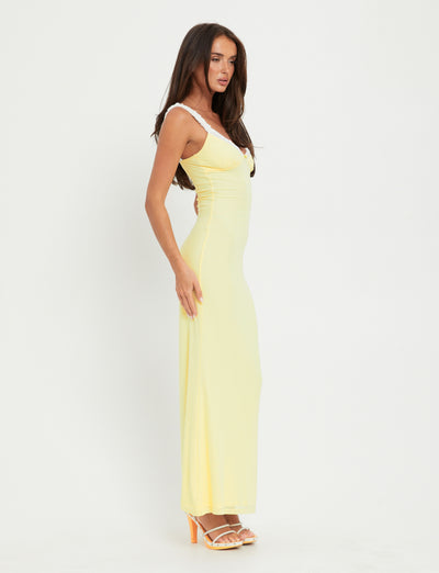 A woman with long hair is standing sideways against a plain white background. She is wearing the sleeveless, floor-length AMARAN DRESS - YELLOW : LEMON by TIGERMIST and white strappy high-heeled sandals. Her left arm is resting along her side while her right arm is slightly bent, resting on her hip.