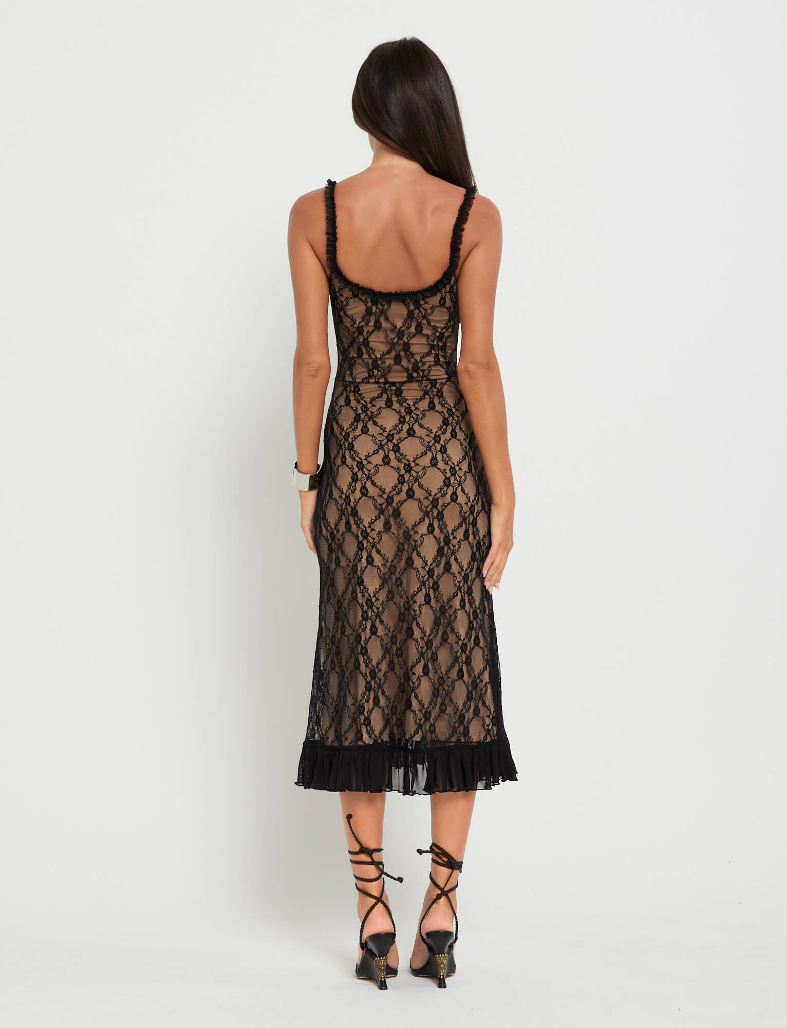 A woman with long brown hair stands facing away, wearing the TIGERMIST DEJA DRESS - BLACK : LACE, a midi-length lace dress with a patterned design and pleated hem, paired with black strappy heels against a plain white background.