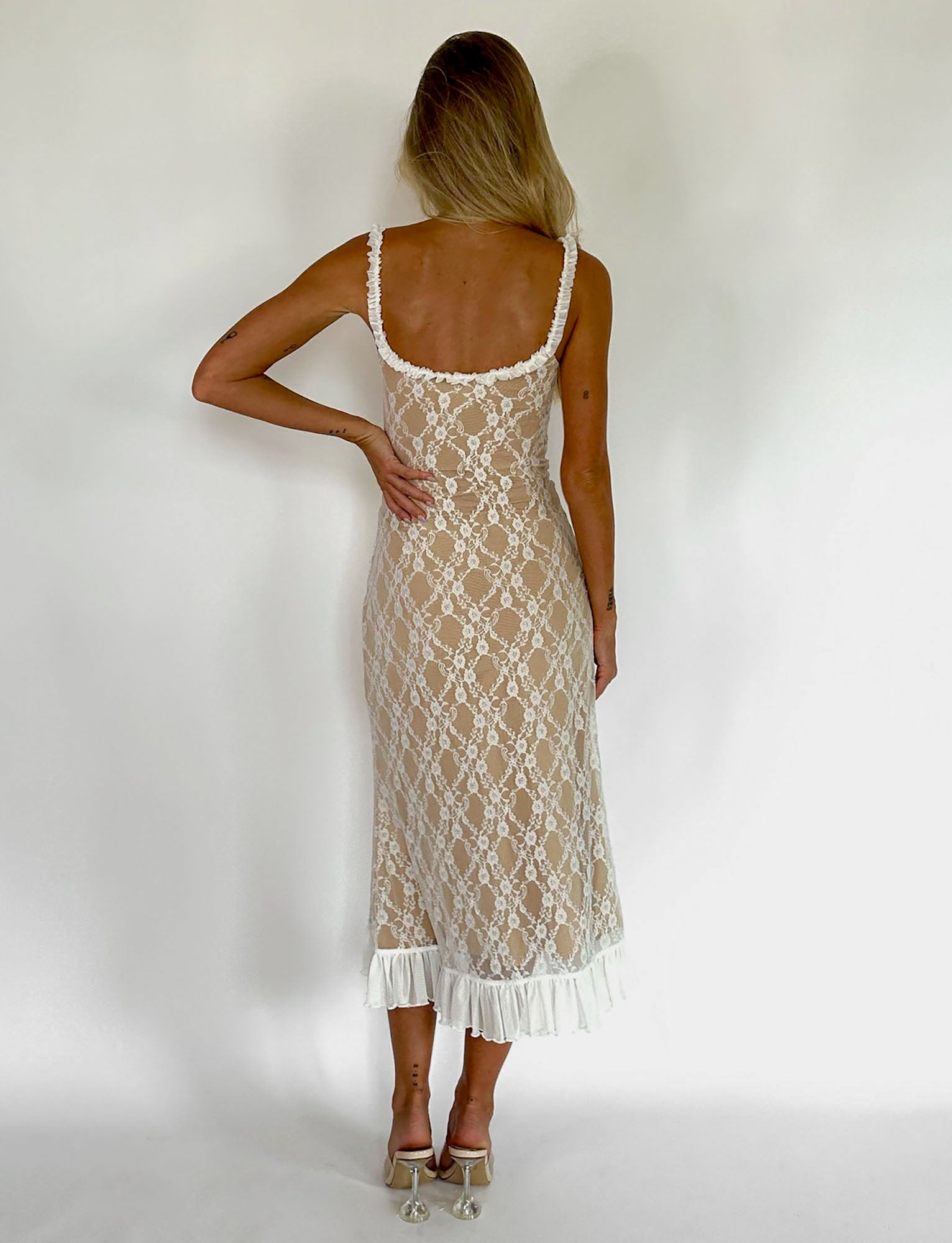 A woman stands with her back to the camera, showcasing the DEJA DRESS - WHITE : LACE from TIGERMIST. The sleeveless, backless white lace dress features a fitted bodice and a ruffled hem. She has long blonde hair and pairs the elegant dress with white high-heeled sandals. The background is plain white.