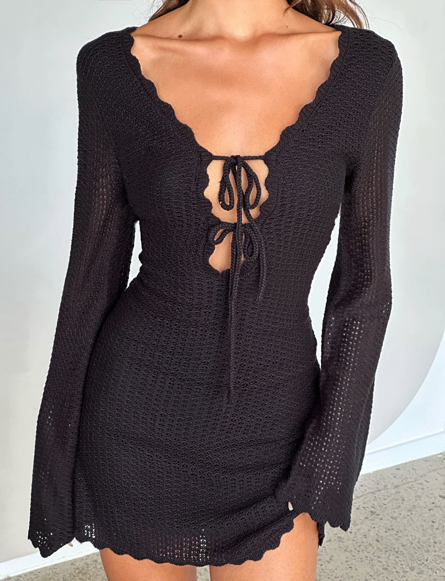A woman is wearing the TEREZA DRESS - BLACK by TIGERMIST. The fitted, long-sleeve crochet dress features a scalloped V-neckline and a tie detail at the chest, allowing a hint of skin to show through the intricate crochet pattern. The background is plain and neutral.