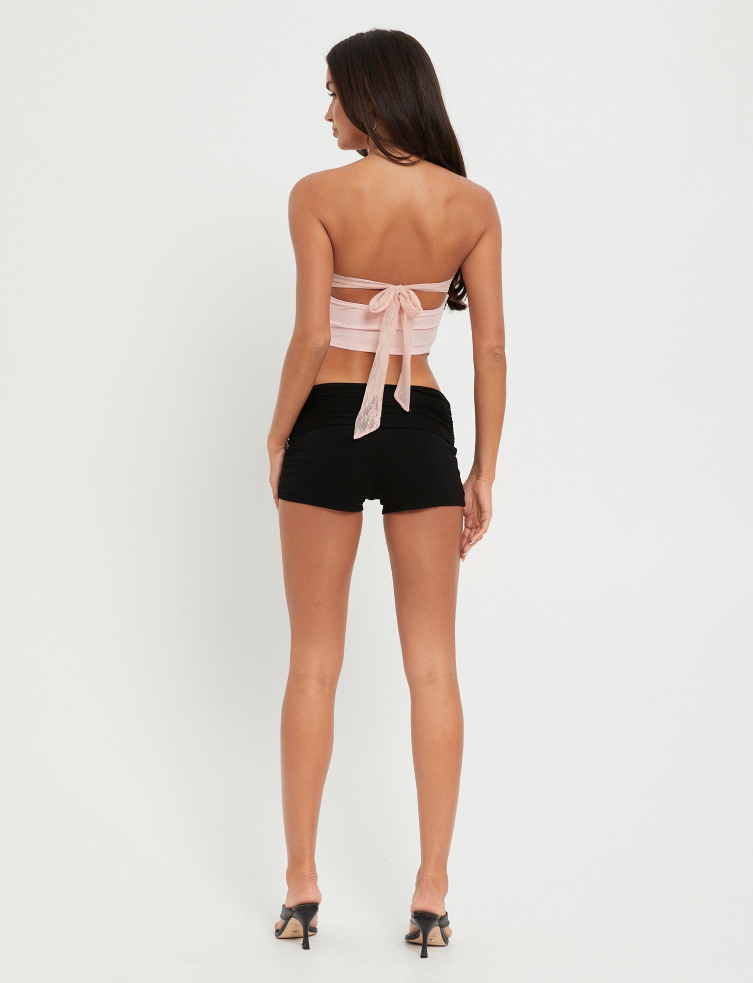 A woman with long hair stands facing away, dressed in the chic HARPER TOP in pastel pink from TIGERMIST, tied with a bow at the back and paired with black shorts and matching high-heeled shoes. The background is plain white.