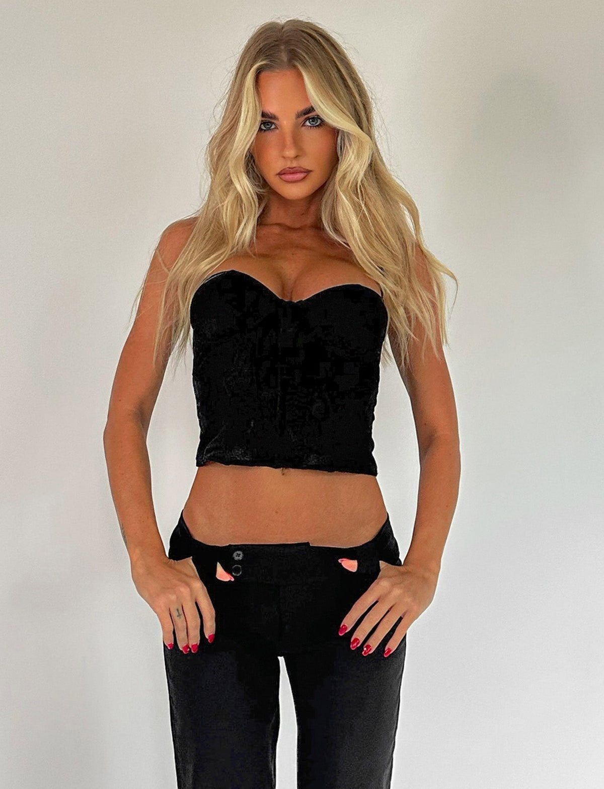 A woman with long blonde hair poses against a white background, wearing the TIGERMIST MAIZIE TOP in black and black pants with side cutouts. She has her hands on her hips and is looking directly at the camera with a neutral expression.