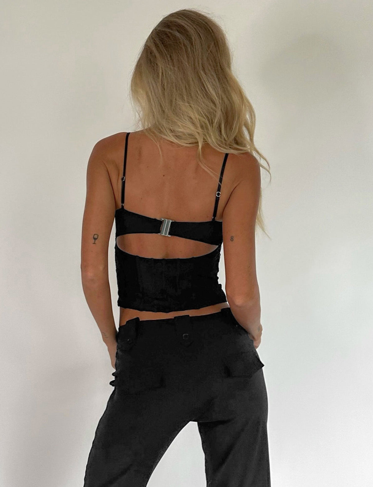 A person with long, wavy blonde hair is facing away from the camera. They're wearing a black MAIZIE TOP by TIGERMIST with a cutout at the back and black high-waisted pants. The background is plain and light-colored.