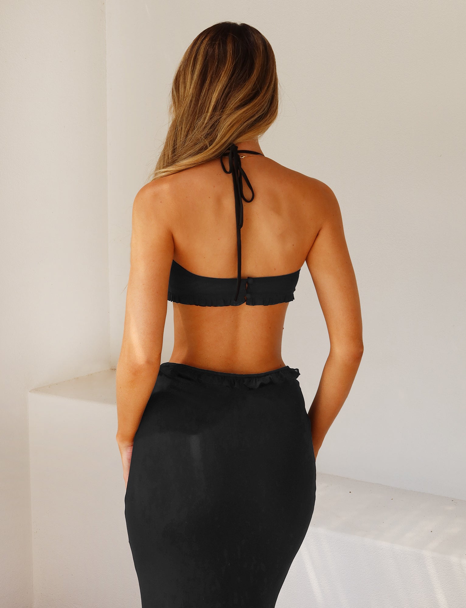 A woman with long, light brown hair stands with her back to the camera, donning the HEAVEN TOP - BLACK and a high-waisted skirt from TIGERMIST. The background is plain and minimalistic, featuring white walls.