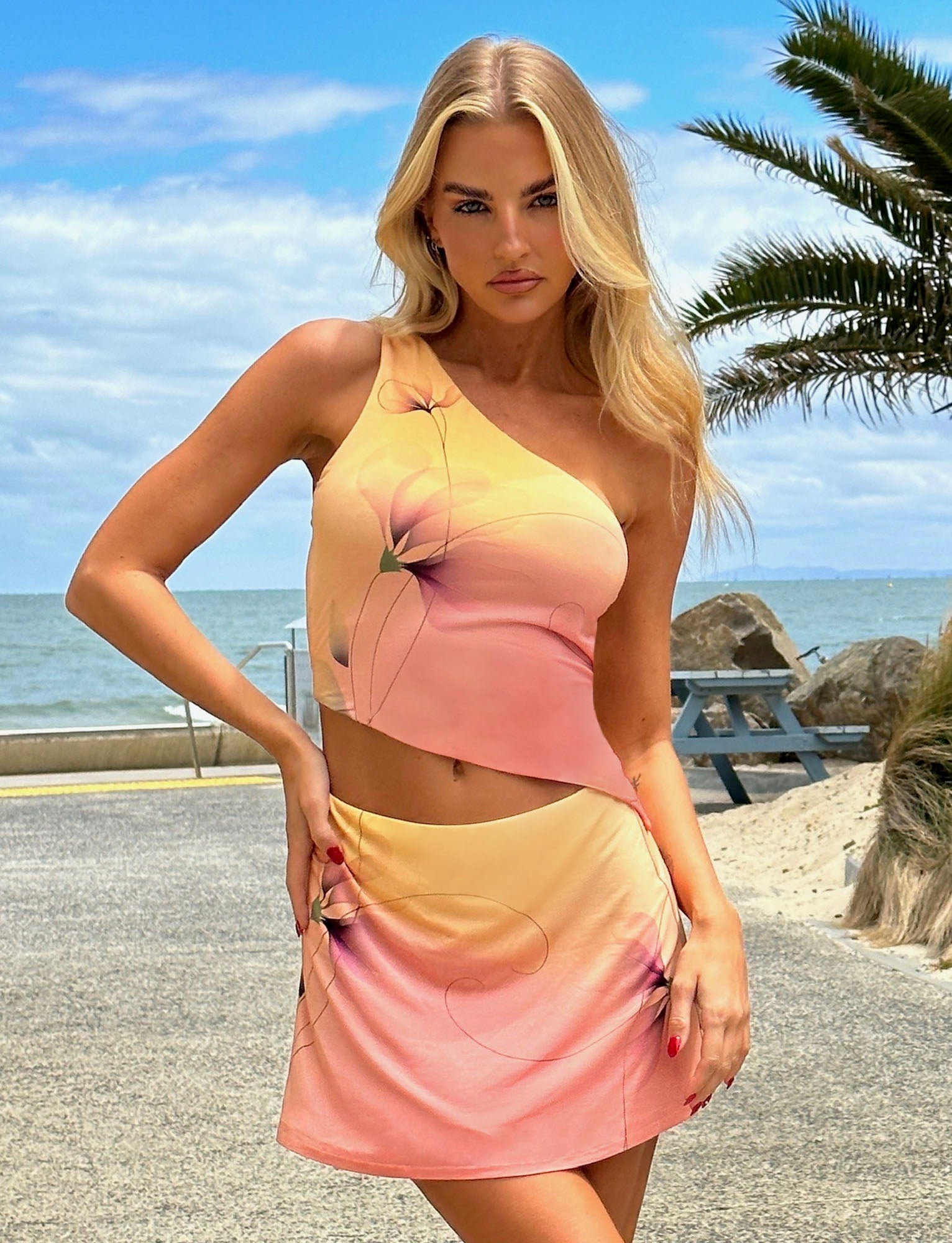 A blonde woman stands outdoors near a beach, with a palm tree and ocean in the background. She is wearing the DREW TOP - MULTI : MACRO FLORAL : GRADIENT FLORAL with a matching mini skirt from TIGERMIST. She strikes a confident pose with her left hand on her hip and her right hand resting on her skirt.