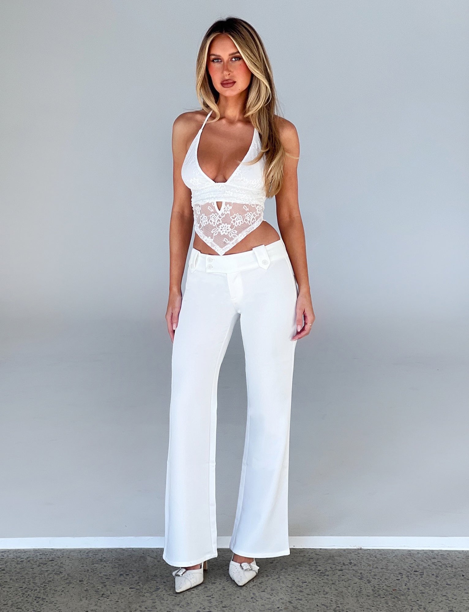 A woman with long blonde hair stands against a plain background, dressed in a chic ensemble by TIGERMIST. She wears the FAYE LACE TOP - WHITE : LACE and high-waisted white pants, paired with white pointed heels. Her neutral expression is directed at the camera, exuding effortless elegance.