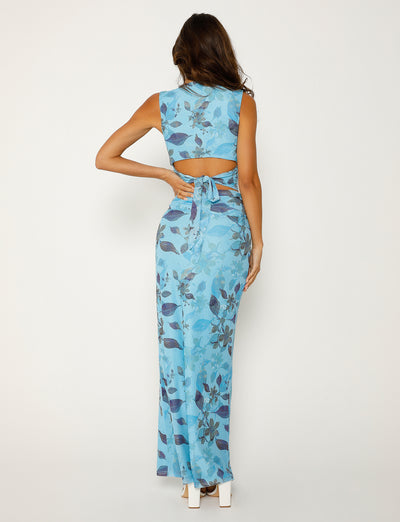 A woman with long wavy hair is facing away from the camera, dressed in a sleeveless Tiger Mist Marietta Skirt - Blue: Watercolour with an open back and a tie detail. The floor-length garment pairs elegantly with her white high heels, as she rests her right hand on her hip.