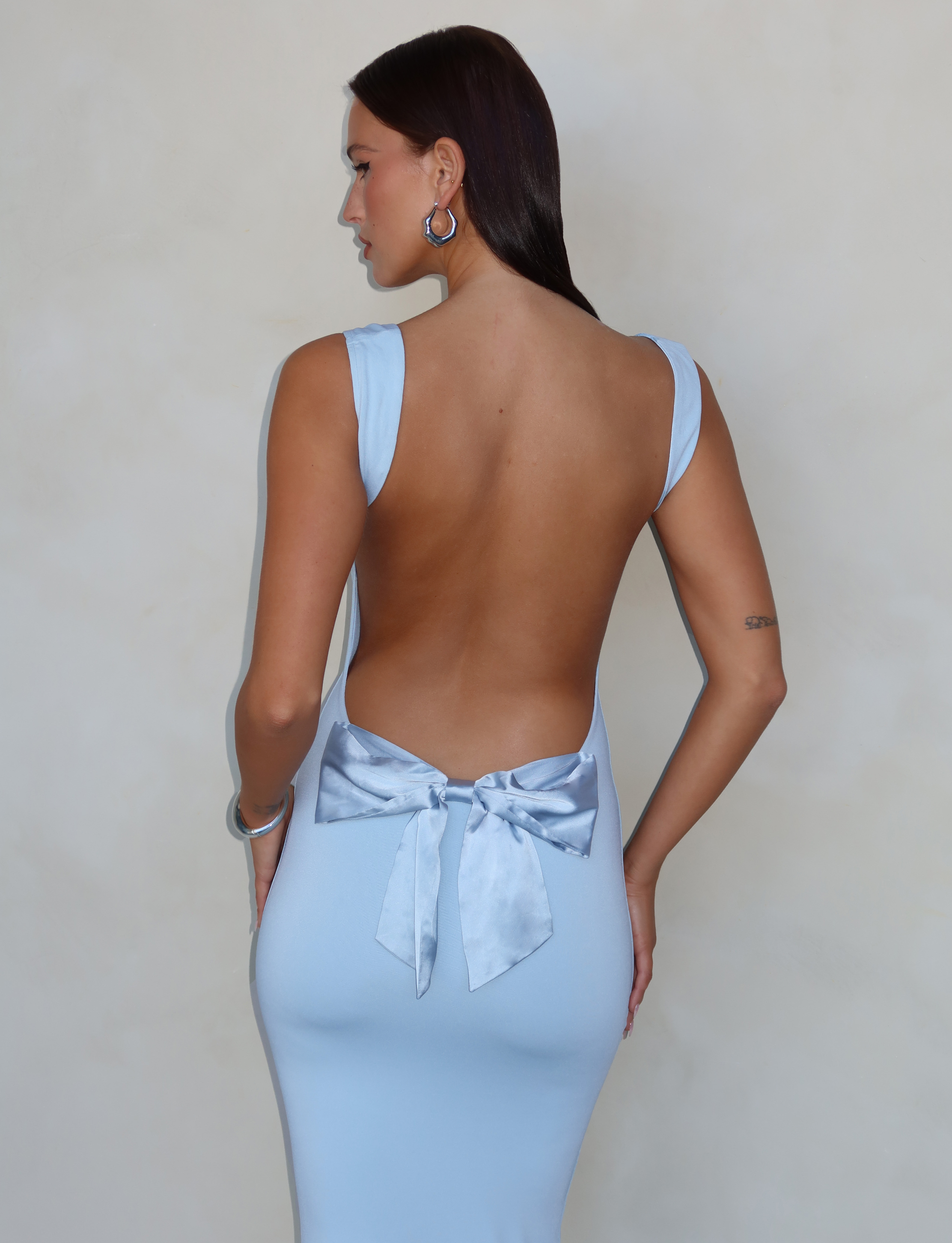 A woman faces away from the camera in the TIGERMIST ZITA DRESS - BLUE: BABY BLUE, showcasing its elegant open back with a satin bow and flattering bodycon silhouette.