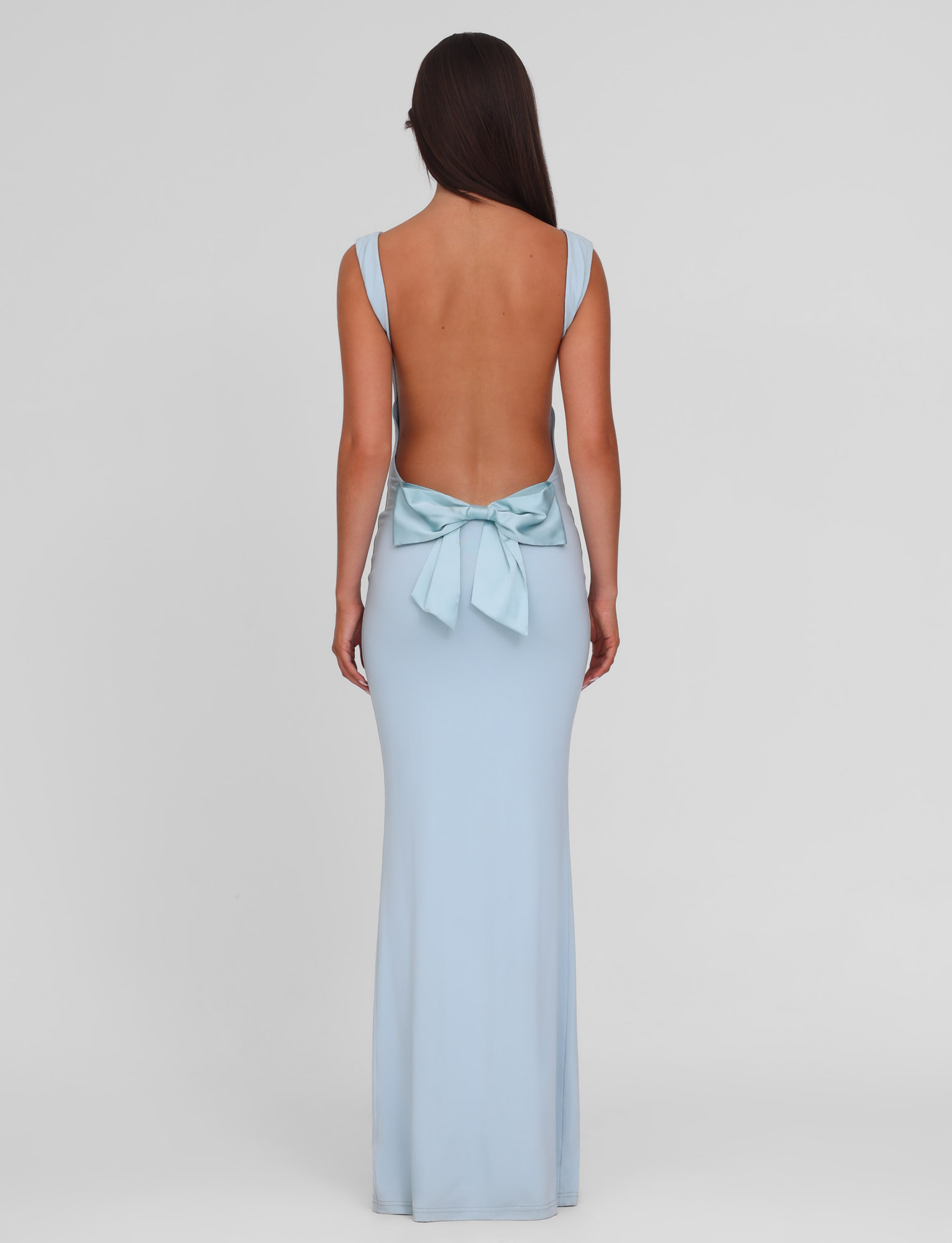 A woman with long brown hair wears the TIGERMIST ZITA DRESS - BLUE : BABY BLUE, a light blue, backless maxi dress with thick shoulder straps and a large bow at the lower back, standing against a plain white background.