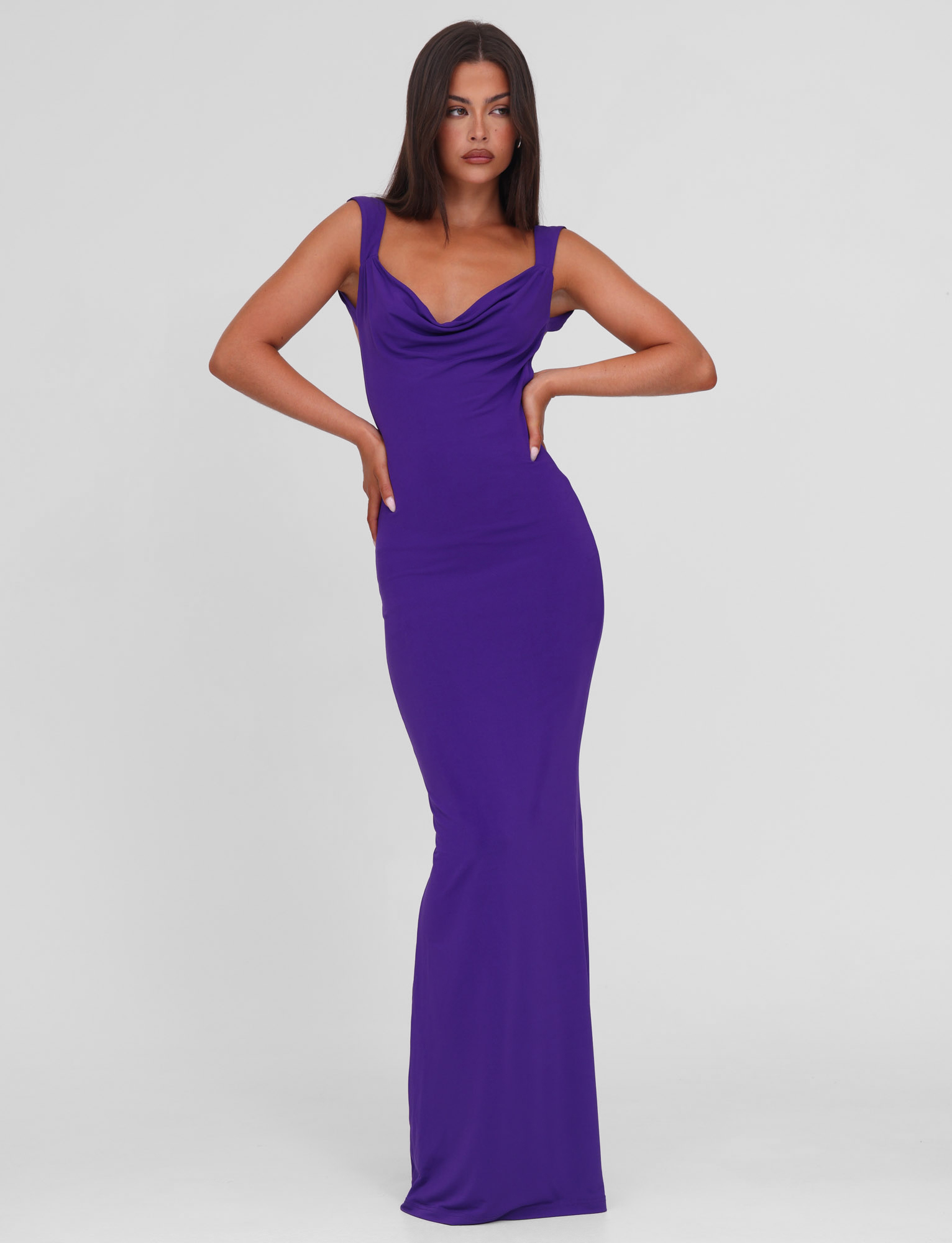 A woman stands confidently in the TIGERMIST ZITA MAXI DRESS - PURPLE, a sleeveless, floor-length bodycon dress with a draped neckline, set against a plain white background.