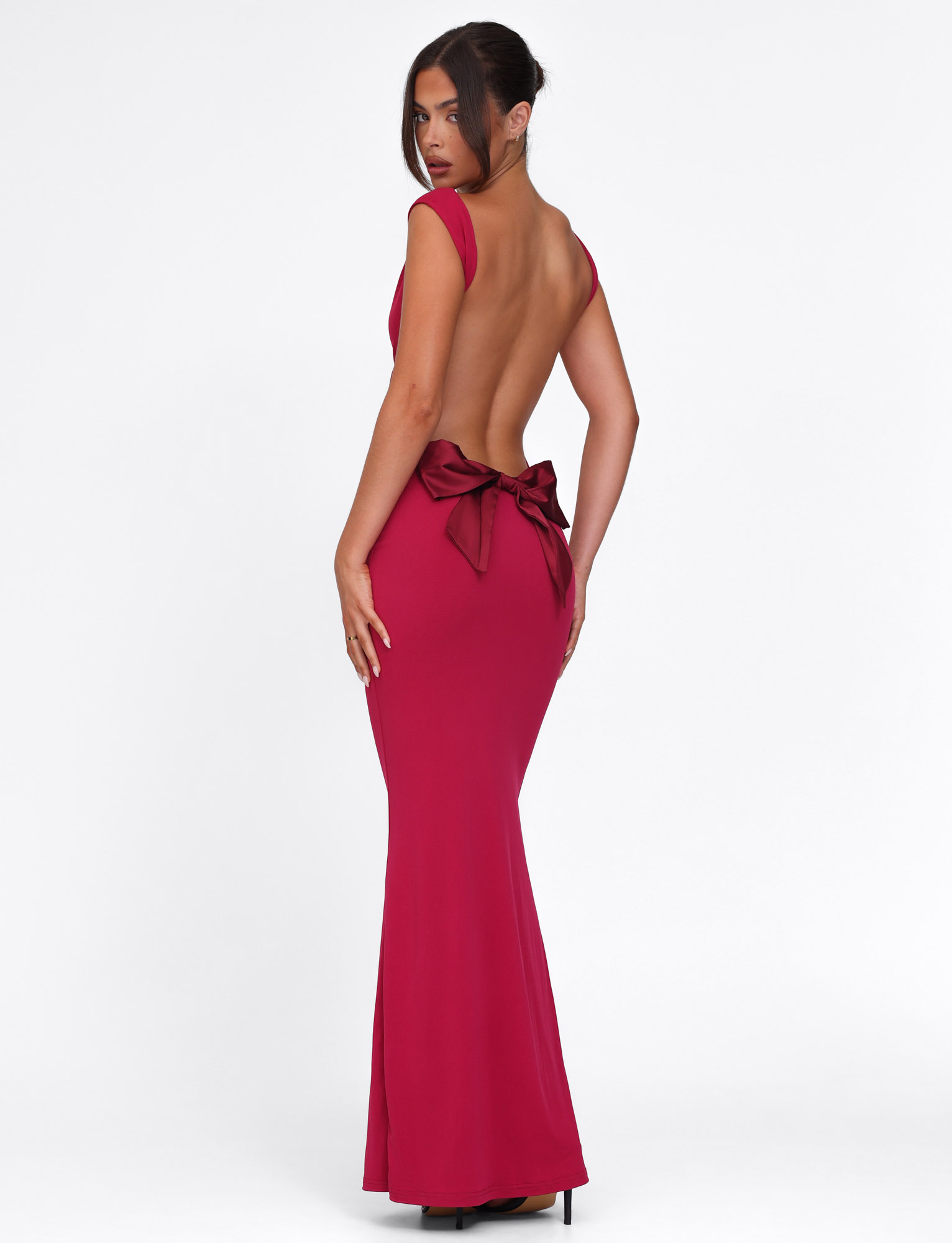 A woman in a sleeveless, backless TIGERMIST ZITA DRESS - RED : SALSA : DEEP RED with a floor-length bodycon fit and bold satin bow at the lower back stands against a white background, glancing over her shoulder.