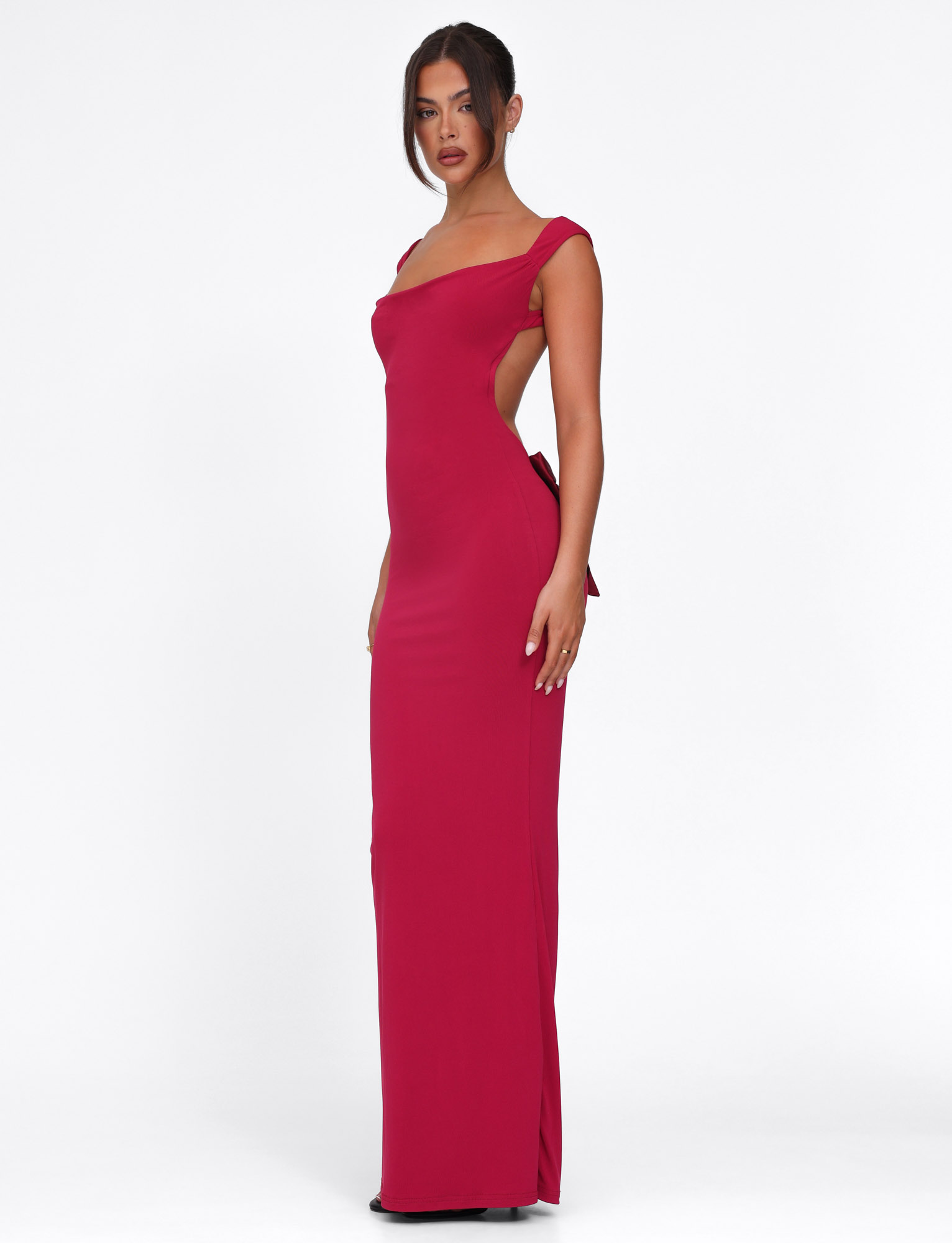 A woman wears the TIGERMIST ZITA DRESS in Salsa Deep Red, a fitted maxi with an open back and side-tie detail. She stands slightly sideways, hair in a sleek updo and her expression neutral.