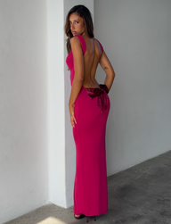 A woman with long brown hair stands indoors on a concrete floor, looking over her shoulder at the camera while wearing the TIGERMIST ZITA DRESS in Salsa (Deep Red), a long, sleeveless, backless dress with a large bow at the lower back.