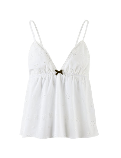 The MILANI TOP - WHITE from TIGERMIST is a white spaghetti strap camisole featuring a subtle floral eyelet pattern. It boasts a V-neckline, gathered detailing under the bust, and a small decorative brown bow at the center front. The fabric appears lightweight and airy.