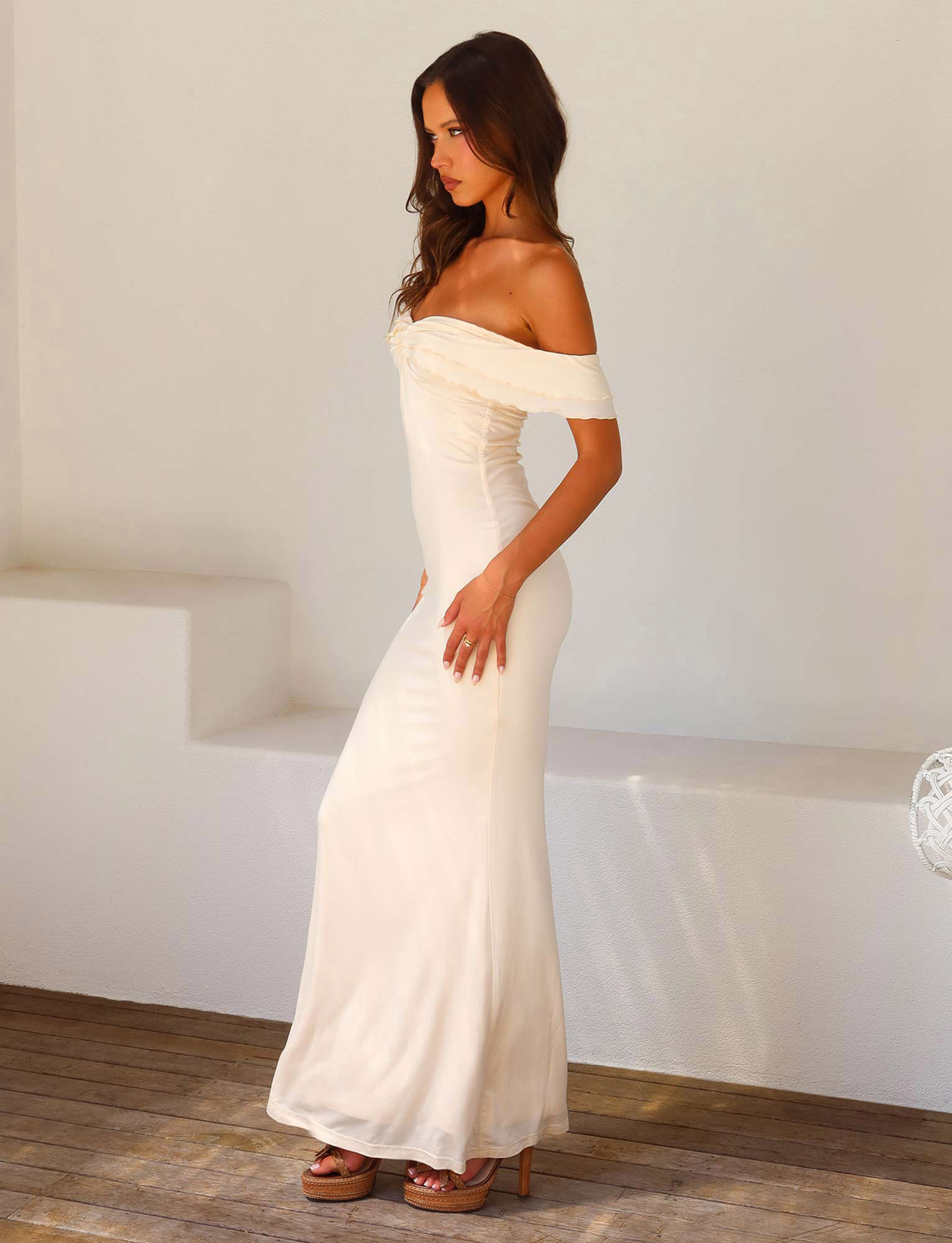 A woman with long brown hair stands indoors against a minimal white wall and wooden floor, wearing the TIGERMIST DENALI MAXI DRESS - WHITE : BONE, a bodycon off-the-shoulder style, paired with high heels.
