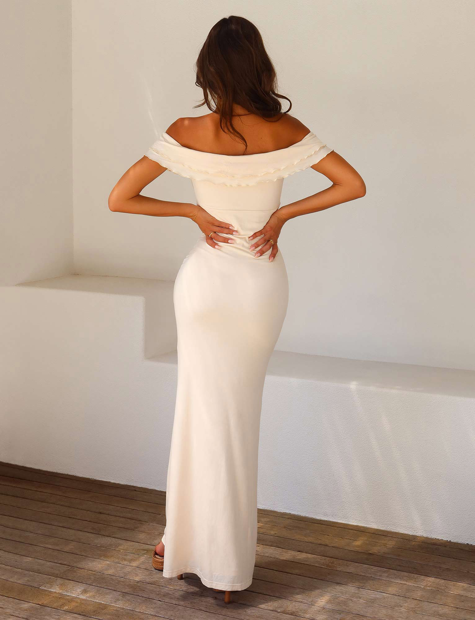 A woman wearing the TIGERMIST DENALI MAXI DRESS - WHITE : BONE, a bodycon off-the-shoulder floor-length gown, stands indoors on wooden flooring with her back to the camera and hands on her waist.