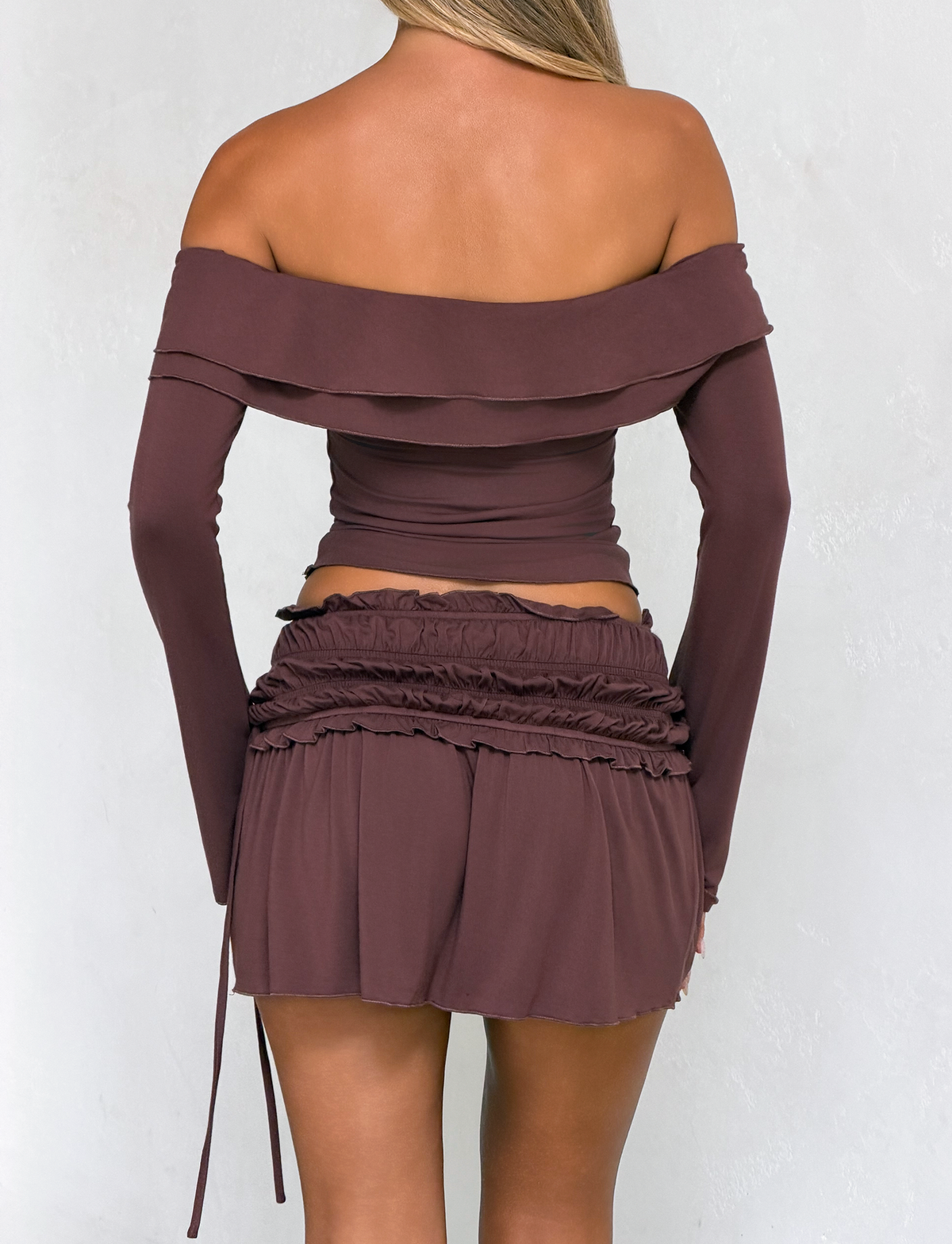 A woman with long blonde hair models the TIGERMIST DENALI MINI SKIRT in chocolate, paired with a matching off-the-shoulder long-sleeve crop top, shown from the back against a plain light background.