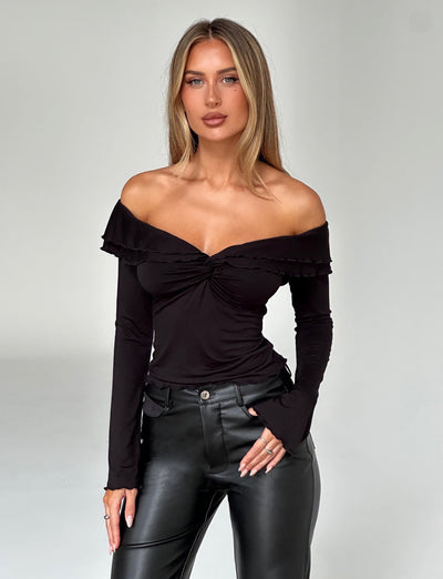 A woman with long blonde hair is standing against a plain white background. She is wearing the DENALI LONG SLEEVE TOP - BLACK from TIGERMIST, which features an off-the-shoulder design with ruffled detailing and long sleeves, paired with black leather pants also from TIGERMIST. She has a neutral expression and her hands are resting by her sides.
