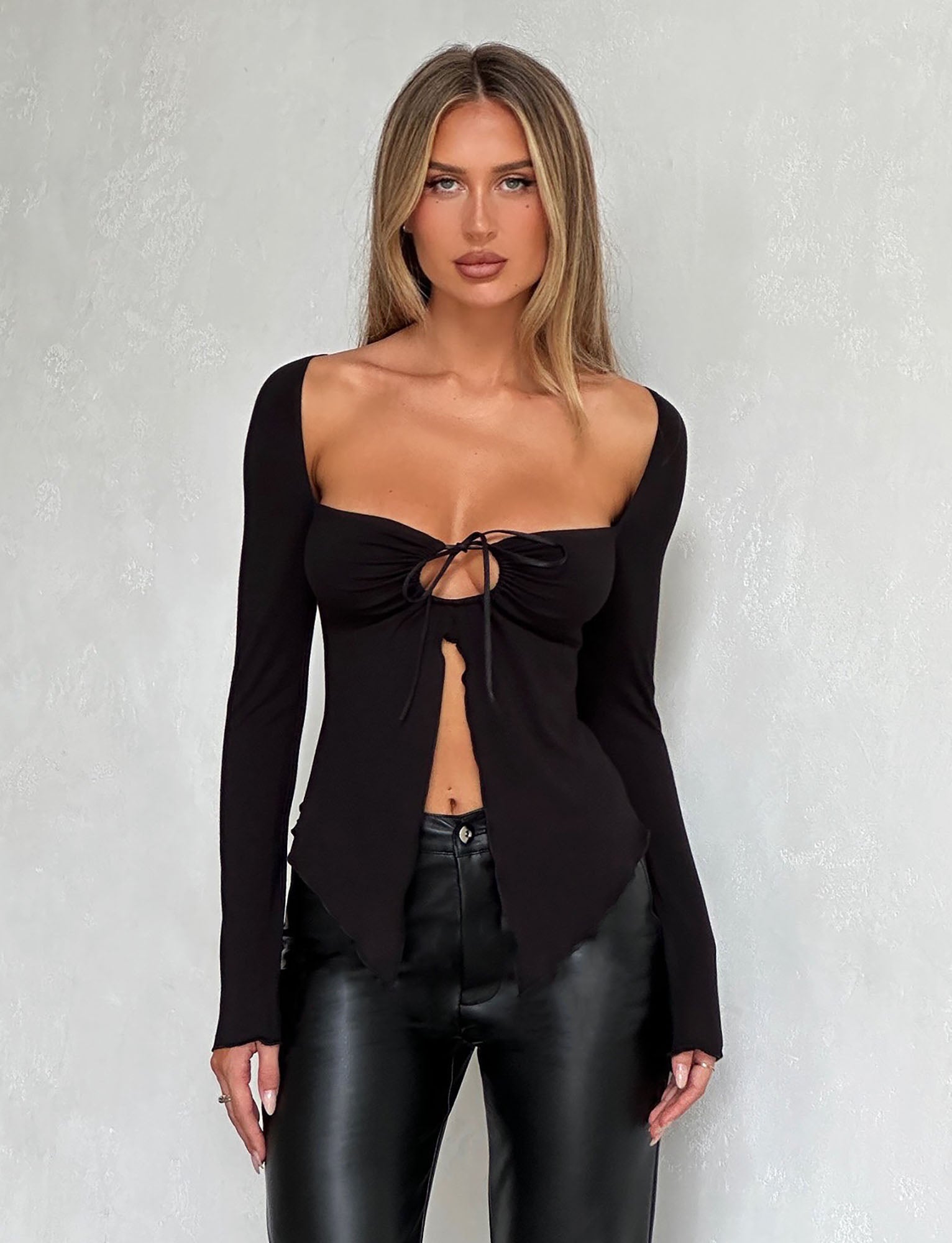 A woman is standing against a light gray textured wall, dressed in TIGERMIST's KAROLINA JERSEY LONG SLEEVE TOP - BLACK and black leather pants. Her blonde hair is styled straight, and she maintains a neutral expression.
