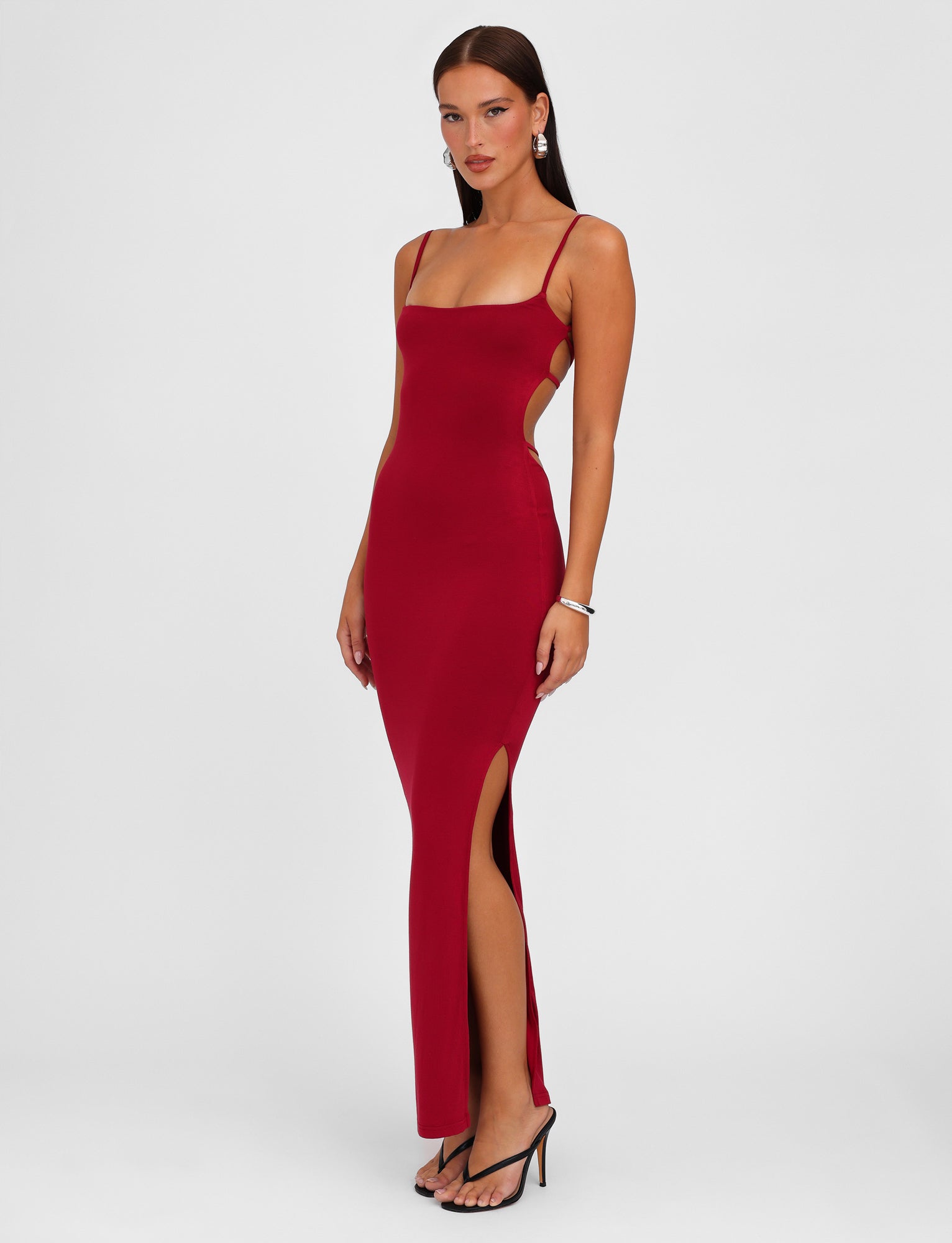 A woman wearing the MINERVA MAXI DRESS - RED : BERRY RED by TIGERMIST, a long, bodycon maxi dress with adjustable shoulder straps and a high slit on one side, stands against a plain white background. She has long hair, wears earrings, and pairs the dress with black high heels. She poses with one hand resting on her side.