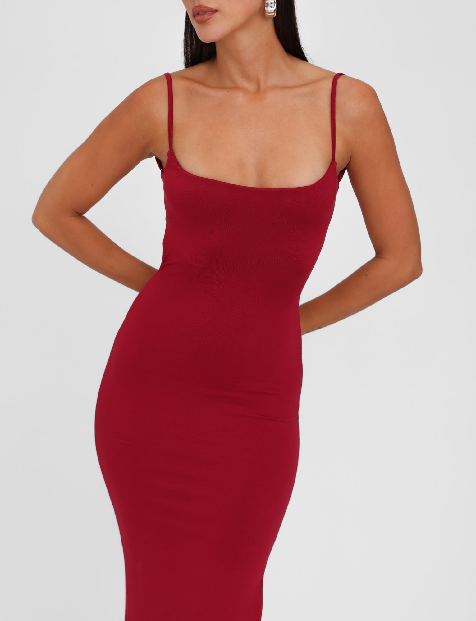 A woman is wearing the MINERVA MAXI DRESS - RED : BERRY RED by TIGERMIST, a sleeveless, form-fitting bodycon maxi dress in a vibrant berry red. The dress features adjustable shoulder straps and a low neckline. Her arms are positioned behind her back, and she has long brown hair paired with large square earrings. The background is plain white.