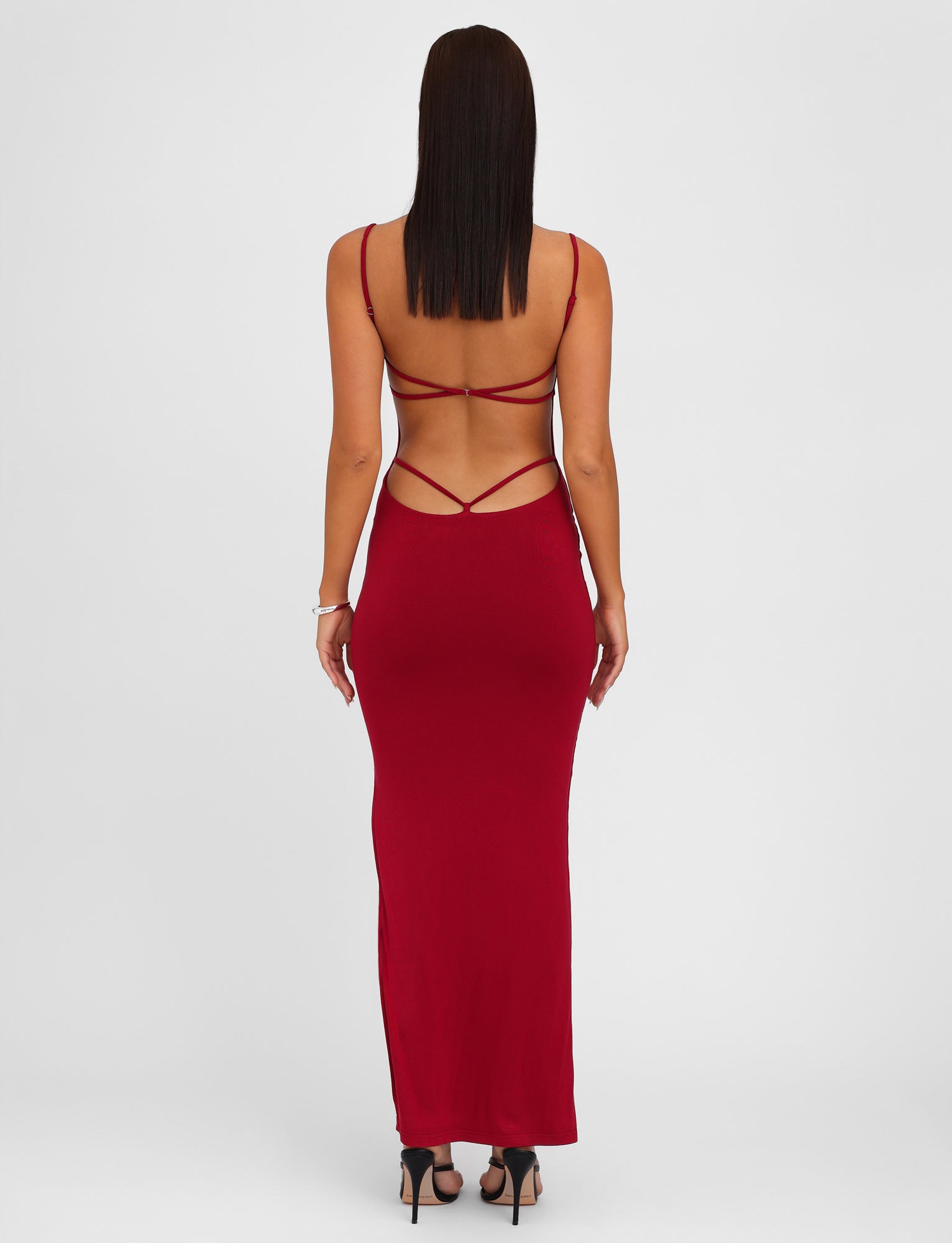 A person stands facing away, wearing a form-fitting, backless MINERVA MAXI DRESS - RED in Berry Red by TIGERMIST. The dress features adjustable shoulder straps and an open lower back design. They have straight, dark hair and are standing in high heels on a plain white background.