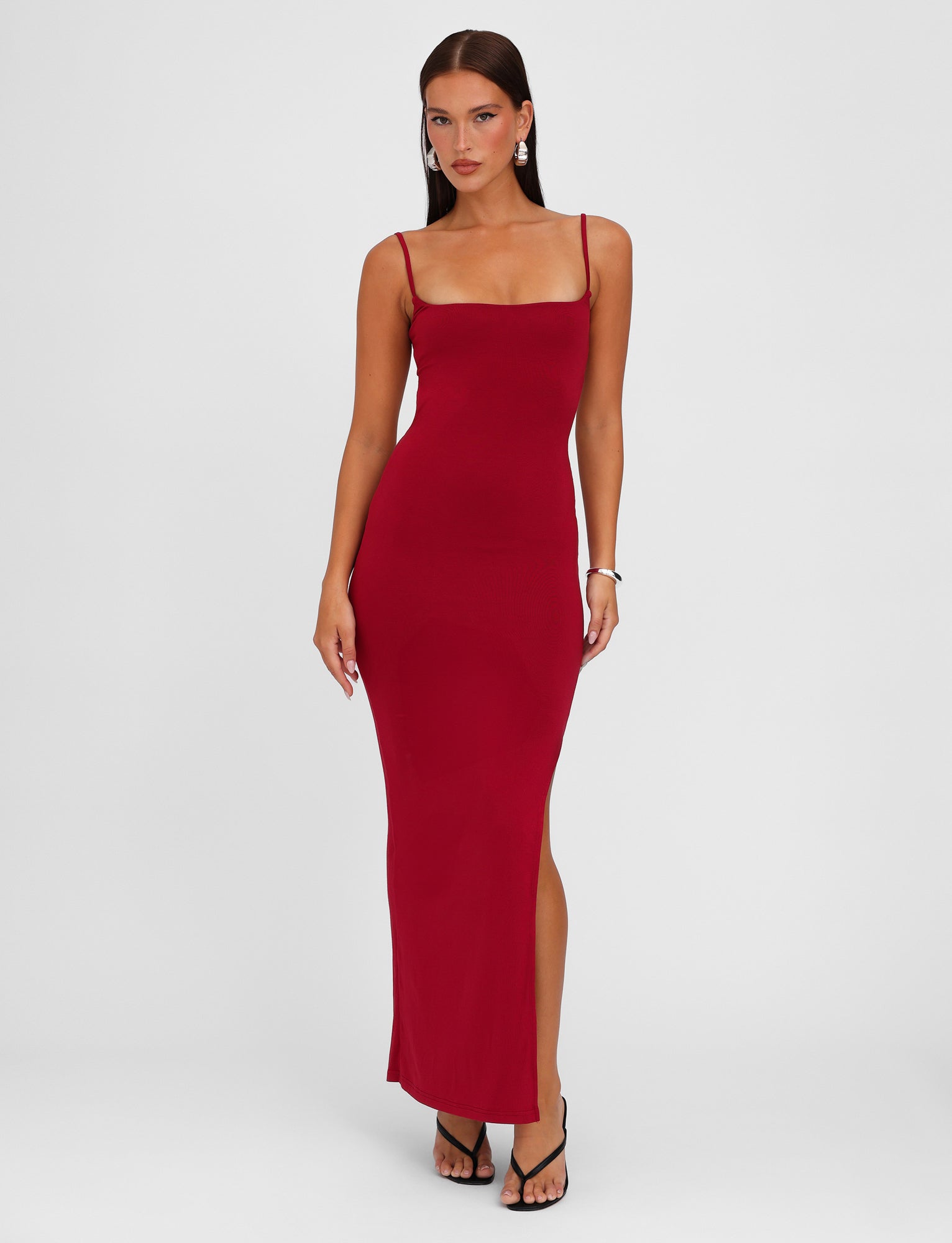 A person stands posing in the Minerva Maxi Dress - Red: Berry Red from TIGERMIST, a long, fitted bodycon maxi dress with adjustable shoulder straps and a side slit. They are wearing black high-heeled sandals and minimal jewelry, with a plain white background emphasizing the outfit.