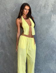 A person with long hair, dressed in a yellow halter top and the LAINEY PANT - LEMON by TIGERMIST, poses against a textured gray wall. They have one hand resting on their thigh while looking off-camera with a serious expression.
