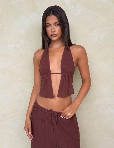 A woman with straight dark hair wears the TIGERMIST LAINEY TOP - CHOCOLATE with matching loose pants, standing against a light beige textured wall and looking at the camera with a neutral expression.