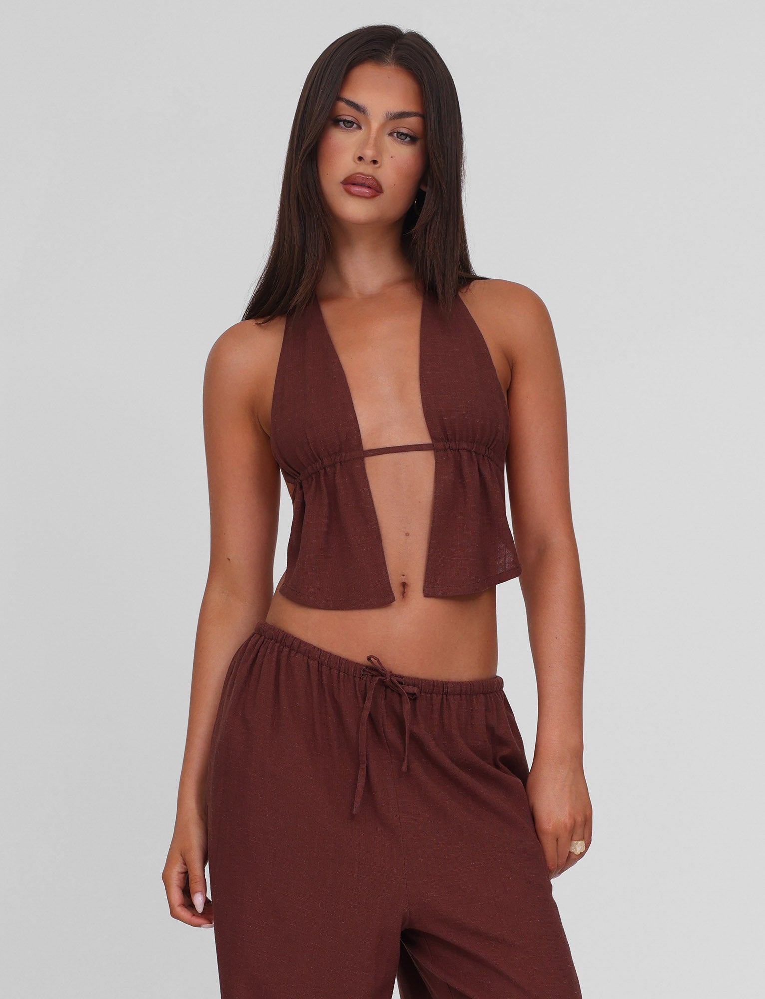 A woman with long dark hair models the TIGERMIST LAINEY TOP - CHOCOLATE, a linen halter top with adjustable ties, paired with matching drawstring pants against a plain light gray background.
