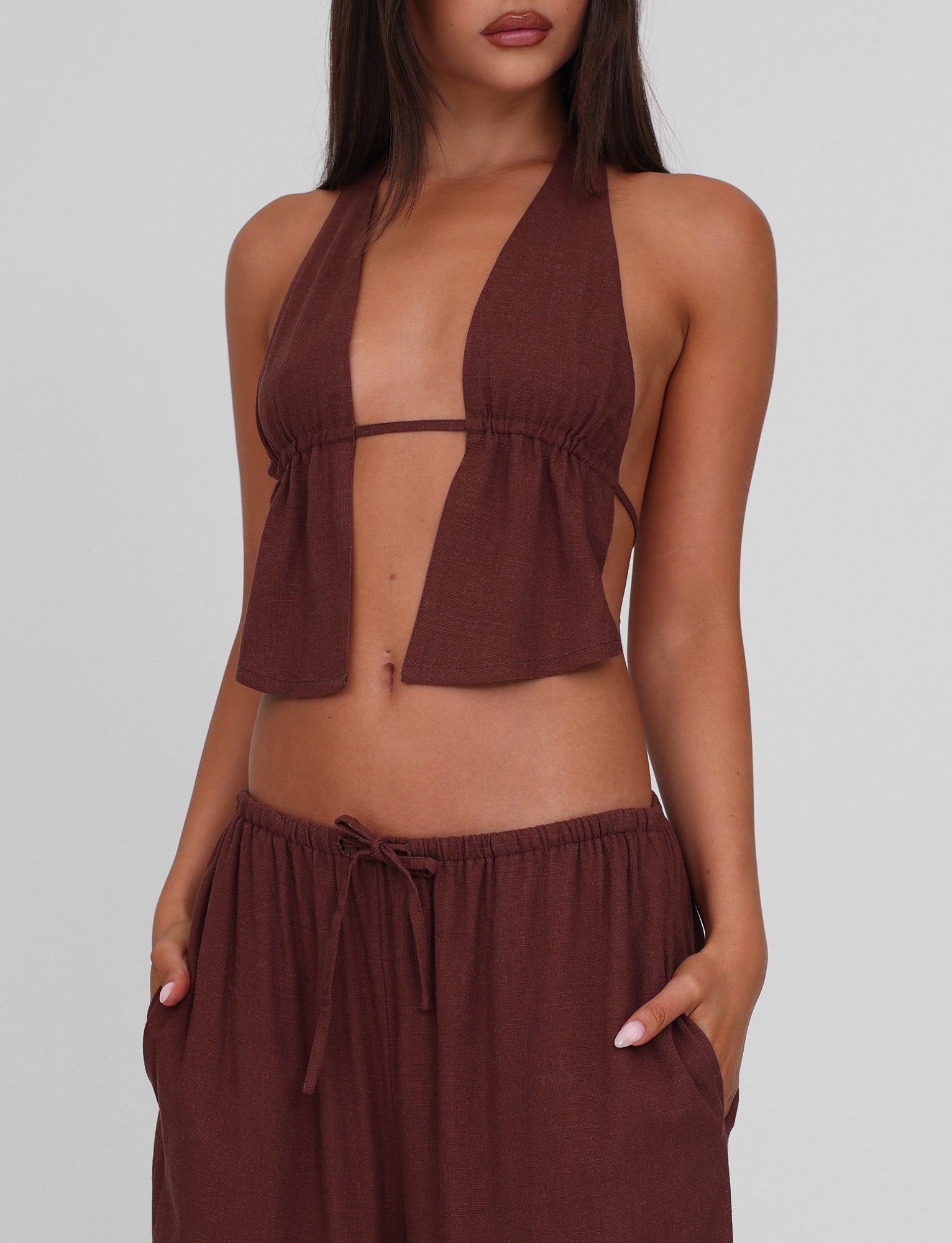 A woman wears the TIGERMIST LAINEY TOP - CHOCOLATE, a sleeveless linen halter with adjustable ties and open front, paired with matching loose drawstring pants. Her straight dark hair contrasts against a light gray background, hands in pockets.
