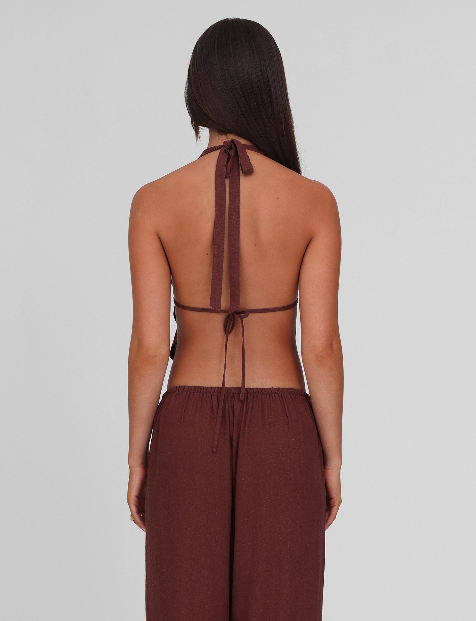 A woman with long dark hair, facing away, wears the TIGERMIST LAINEY TOP - CHOCOLATE with adjustable ties and matching high-waisted pants against a plain light gray background.