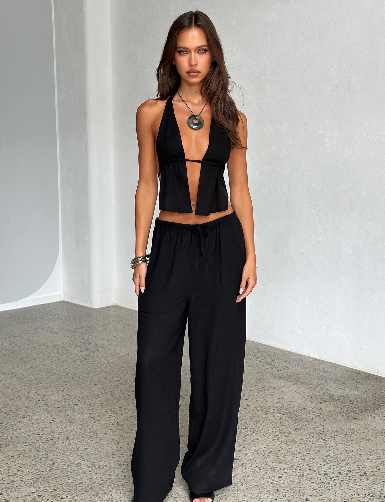 A woman with long brown hair poses against a light gray wall, stunningly dressed in TIGERMIST's black halter top with a deep V-neck and the LAINEY PANT - BLACK. She accessorizes with a large pendant necklace and bracelets on one wrist. The setting appears to be an indoor space with a polished floor.