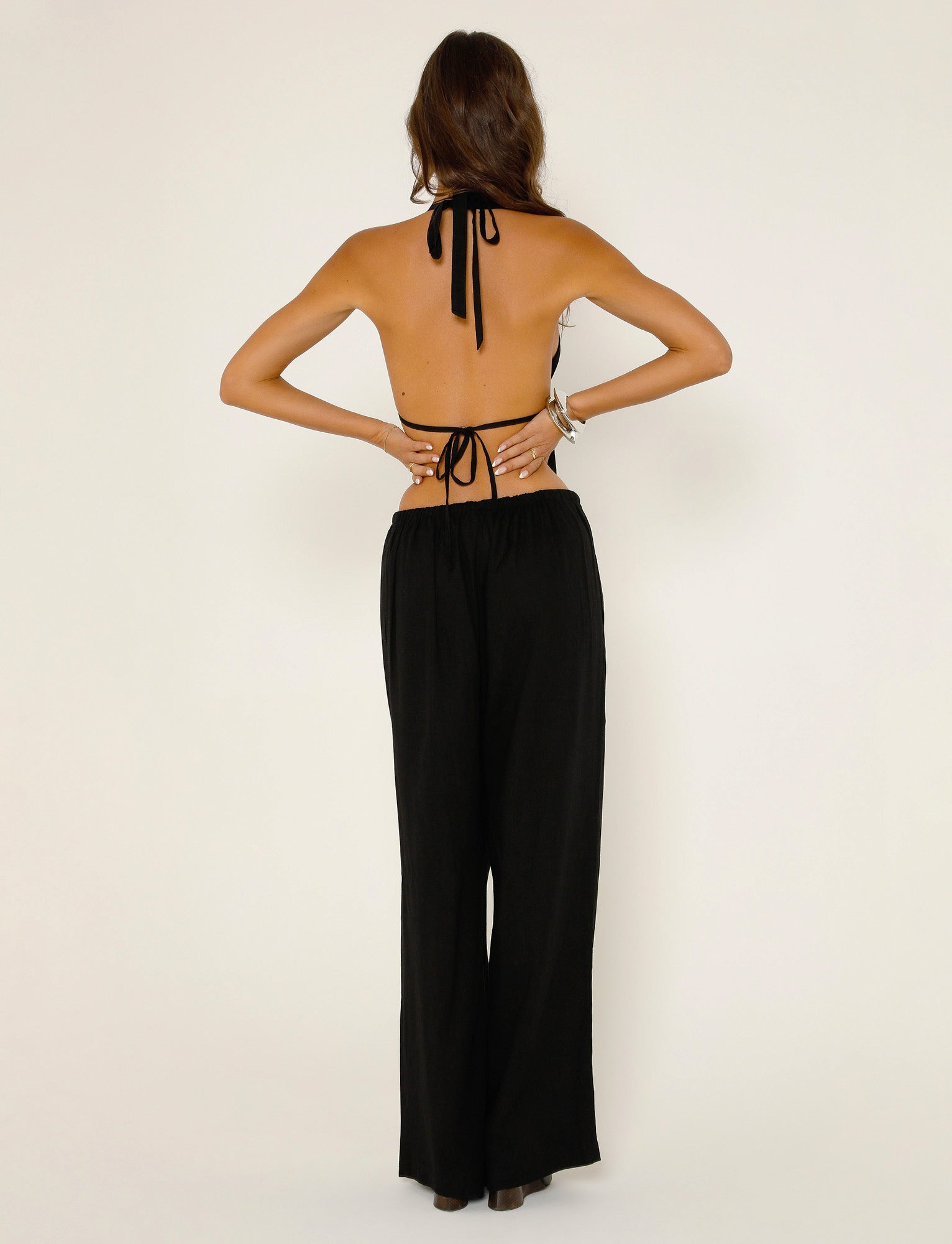 A woman stands facing away from the camera, wearing a halter-neck top from Tiger Mist with thin straps tied at the back and around the neck, paired with TIGERMIST's LAINEY PANT in black. She has long brown hair and is posing with her hands on her hips against a light background.
